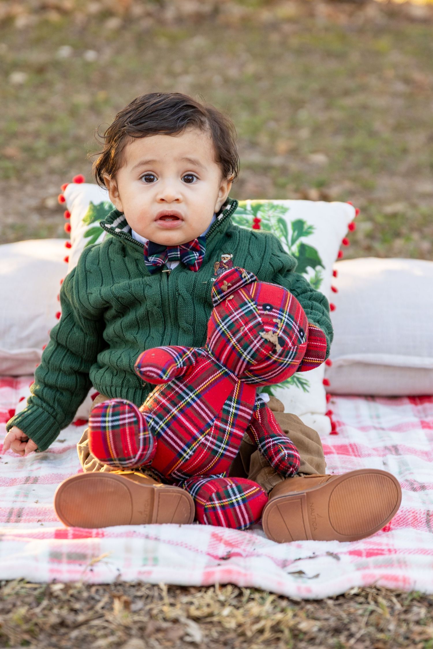 A small child in a green sweater sitting on a plaid blanket holding a red plaid teddy bear.