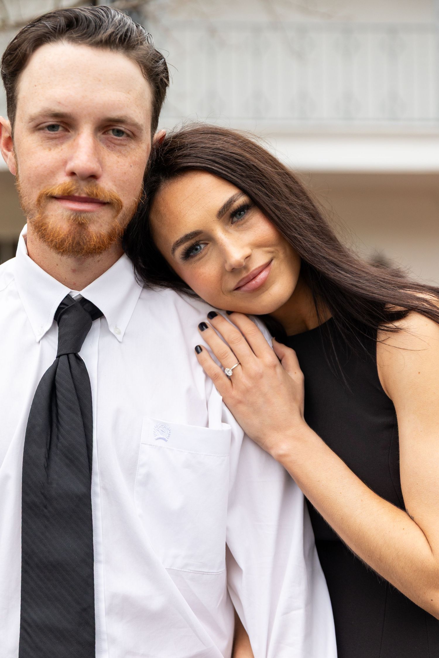 A romantic engagement photo showing a couple embracing, with the woman displaying her ring while wearing a black dress.