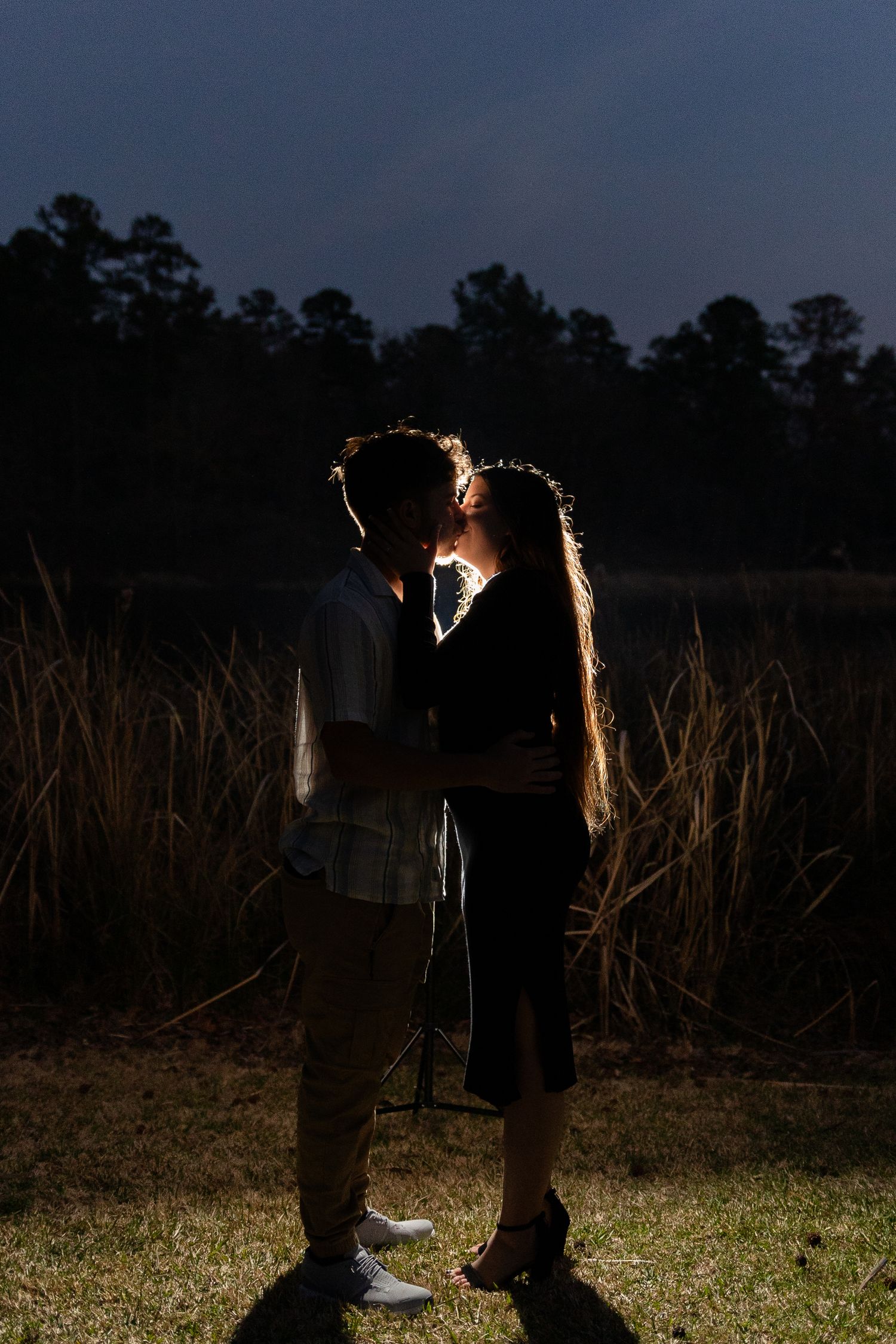 A silhouetted couple stands together in a field at dusk with dramatic backlighting.