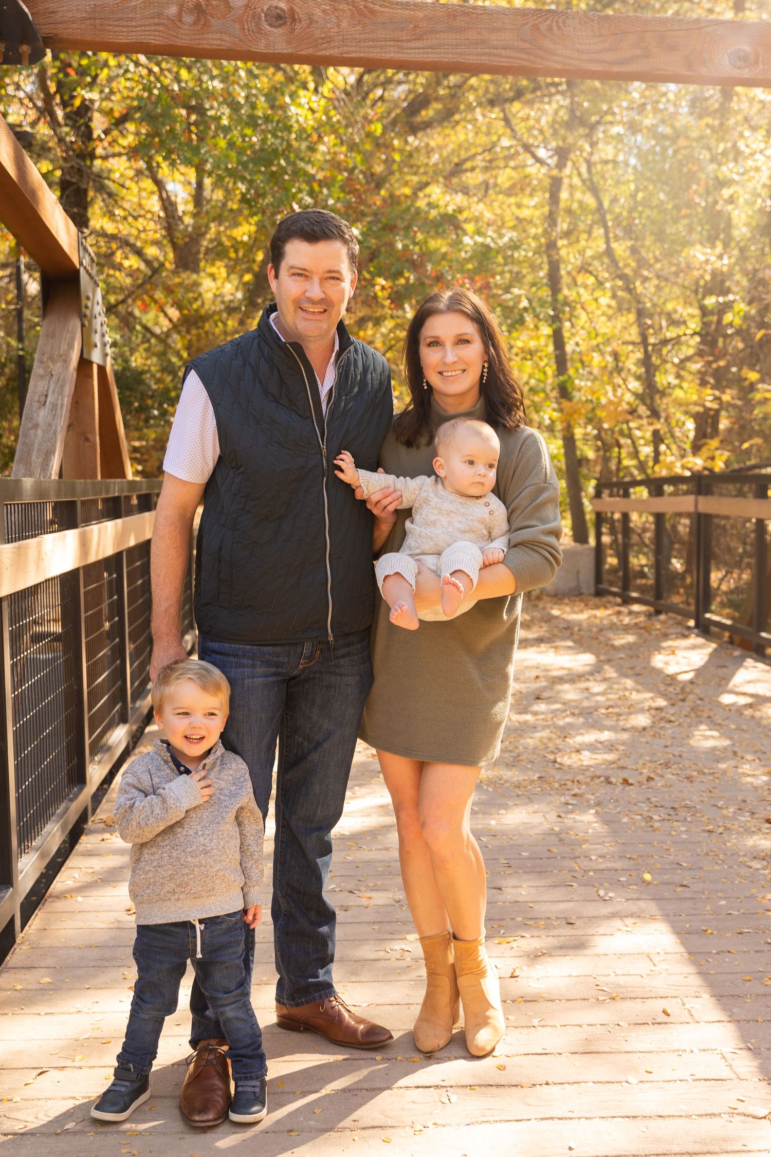 Family of four poses together on wooden bridge path during fall season.