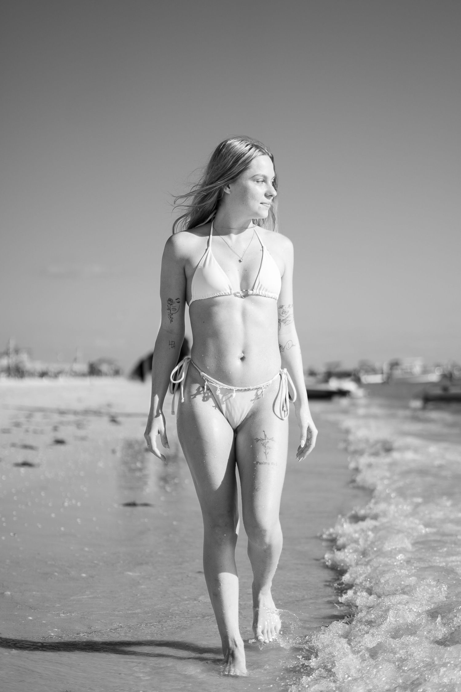 Slender woman in white bikini walking along ocean shoreline in black and white beach photo.