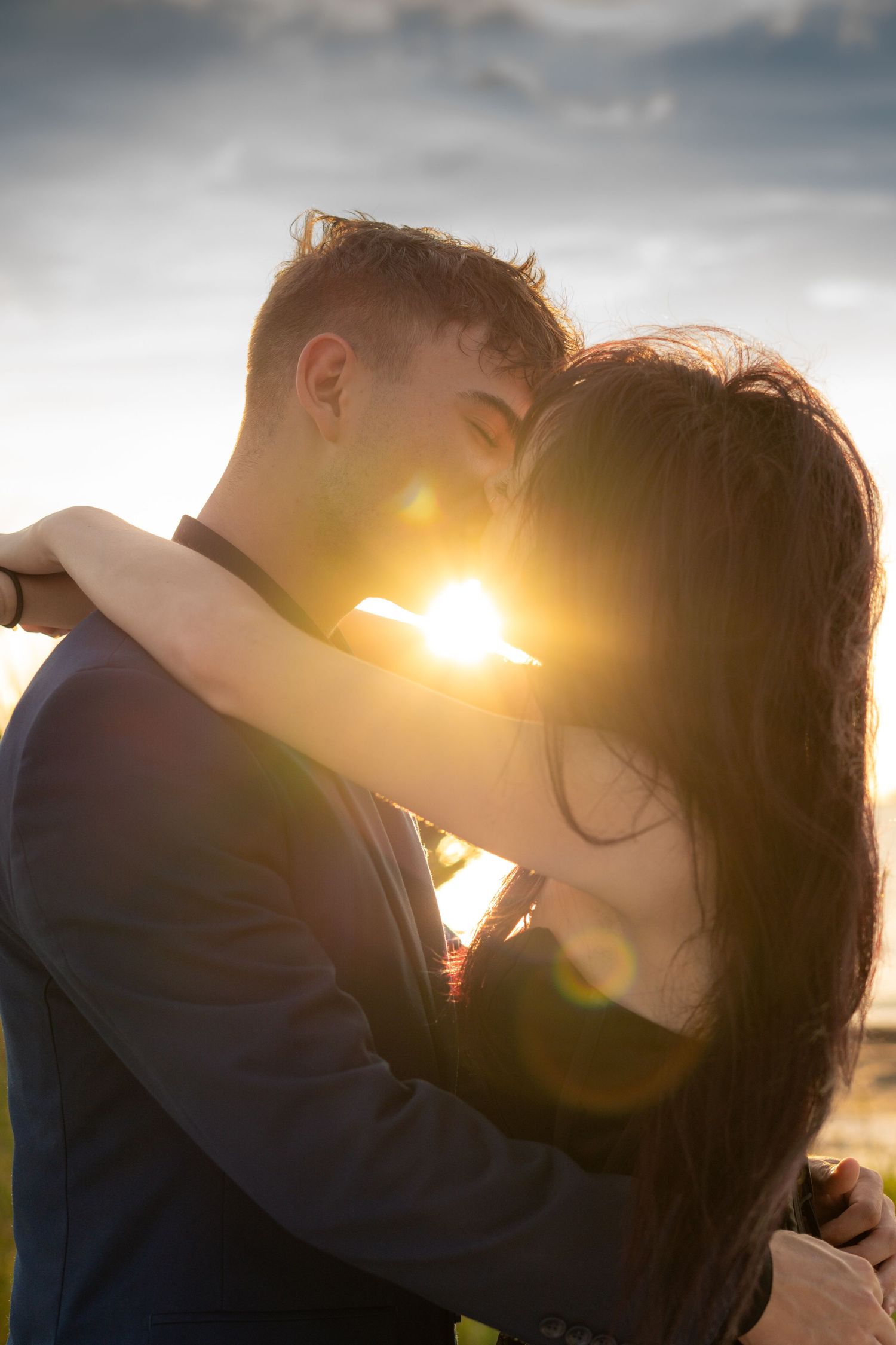 Romantic couple embracing at sunset with golden lens flare between them against a cloudy sky.