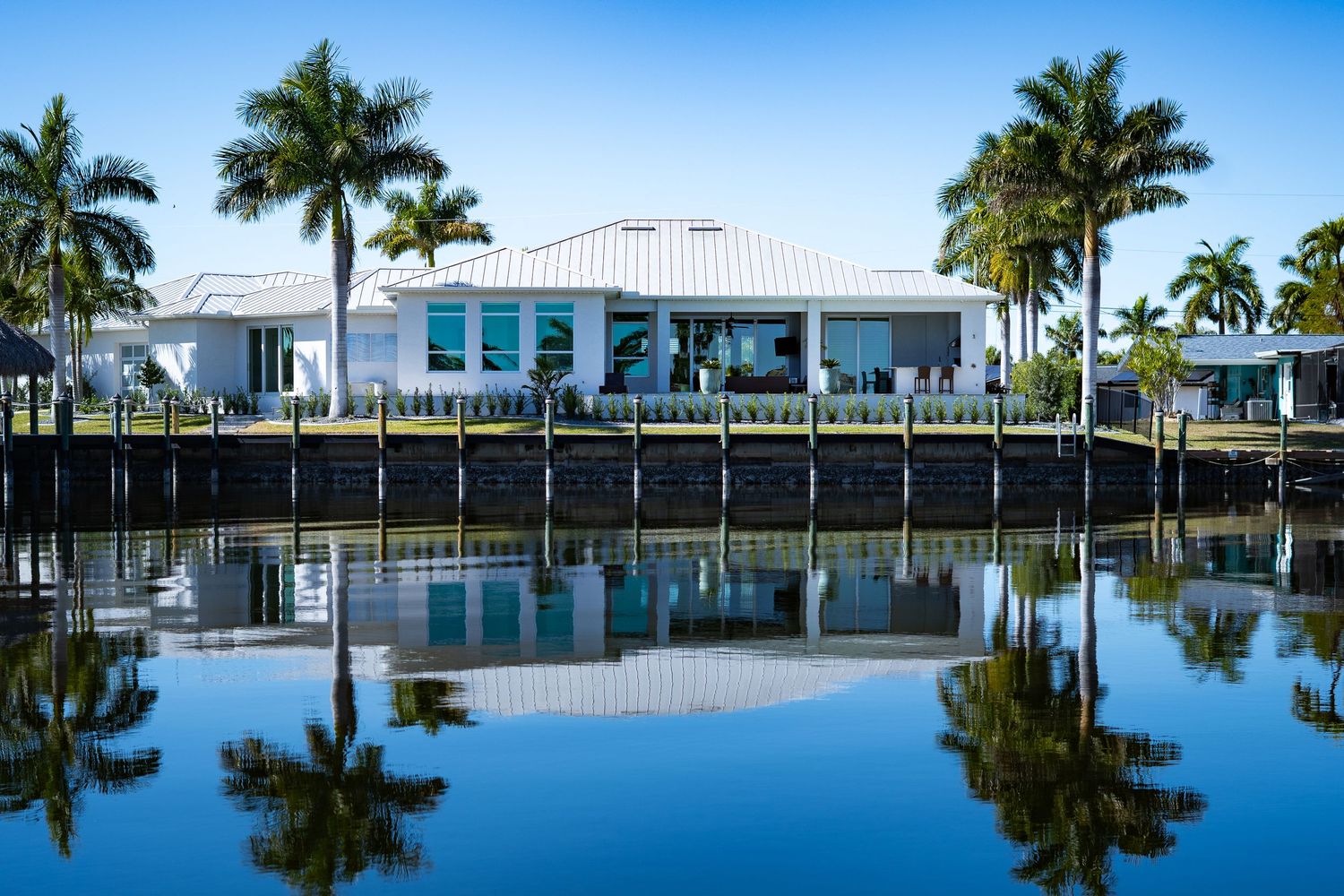 Modern white waterfront home with palm trees reflecting perfectly in calm canal water.