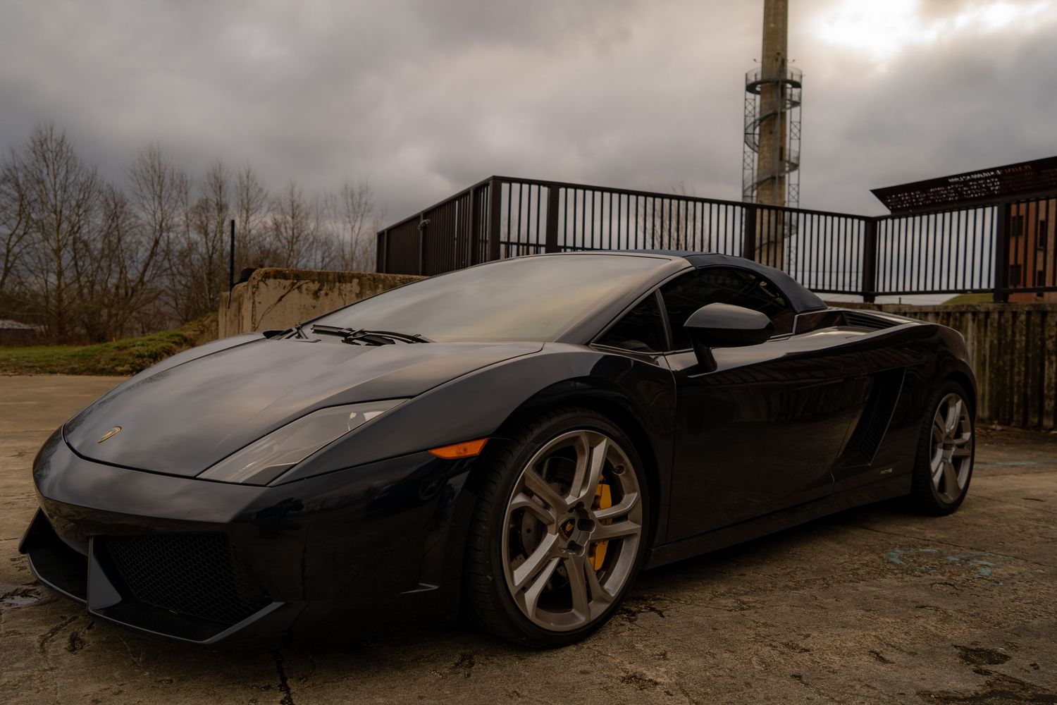 Sleek black Lamborghini Gallardo sports car parked on gravel under dramatic cloudy sky.