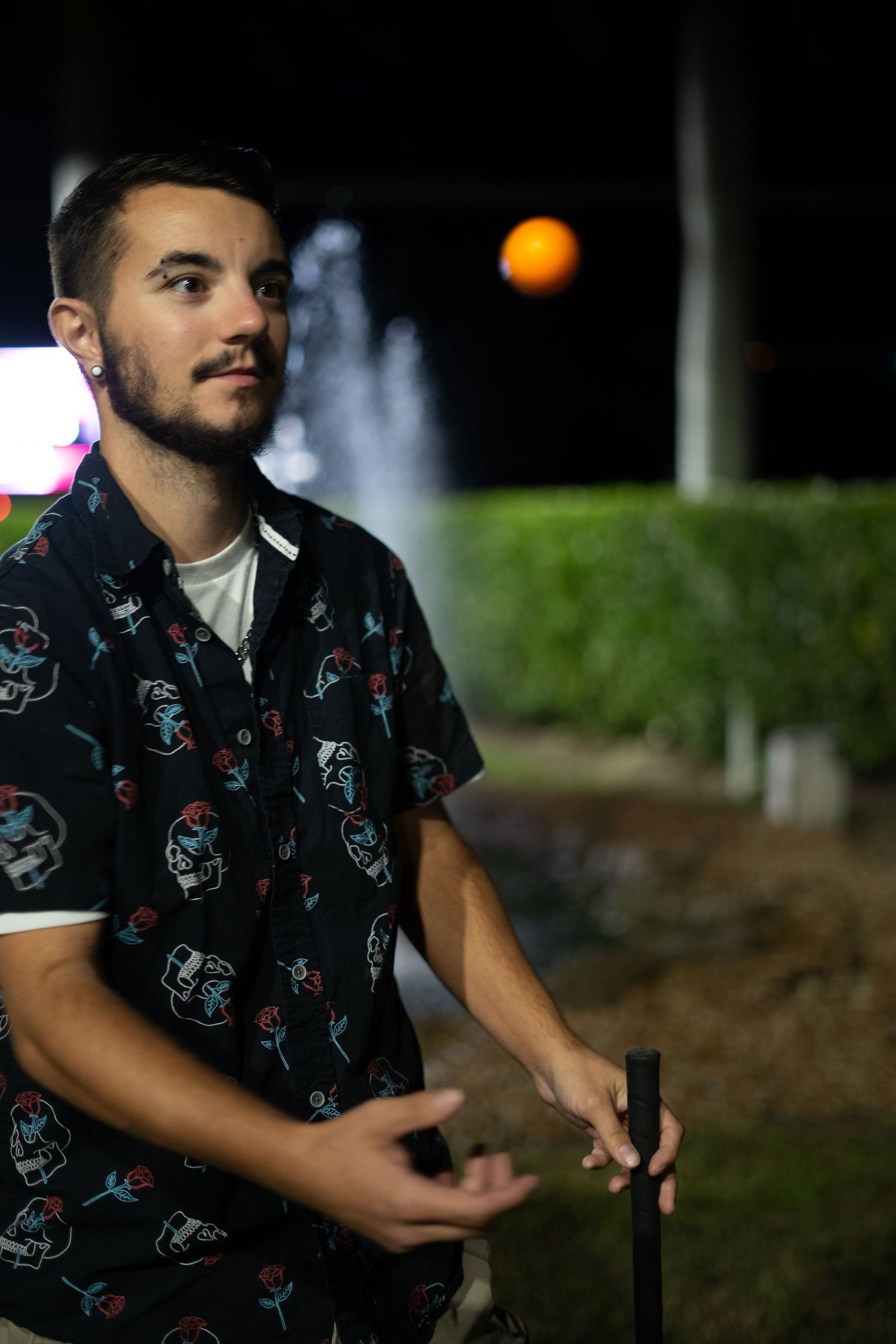 Young man in patterned shirt standing outdoors at night near fountain.