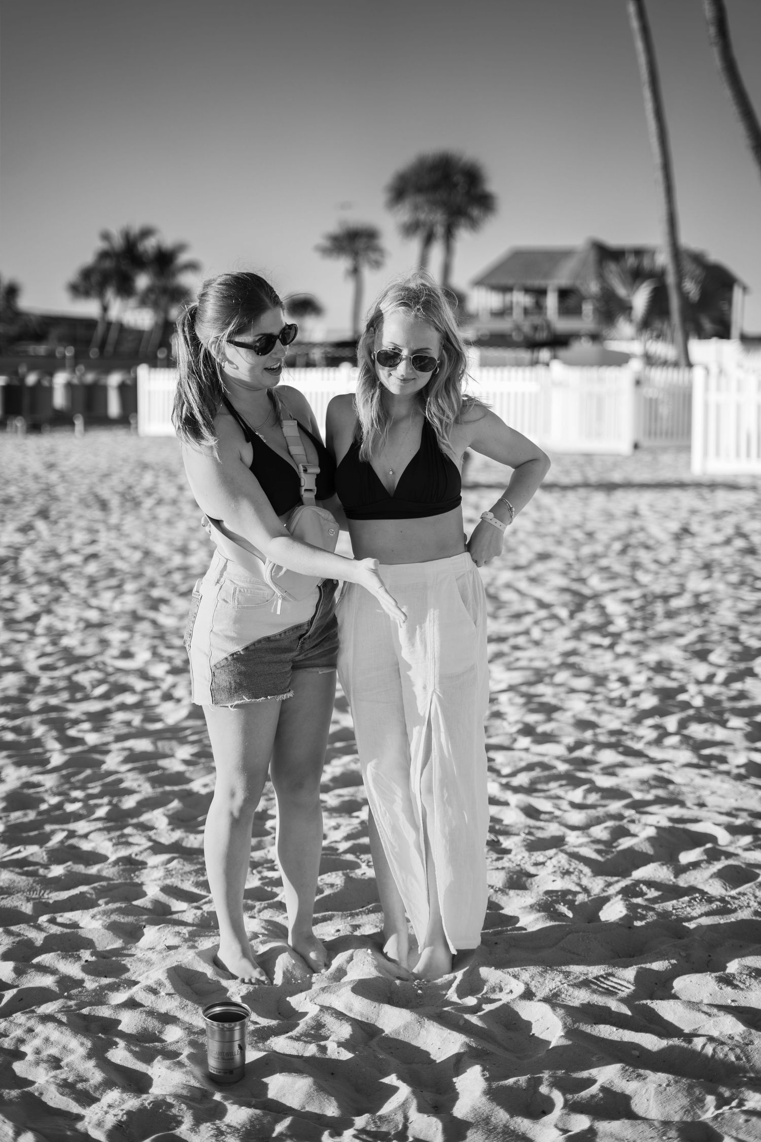 Two women in swimwear and sunglasses standing together on a sandy beach in black and white photo.