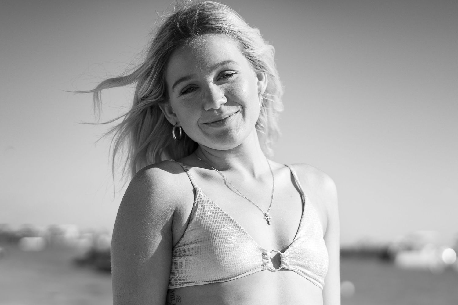 Smiling blonde woman in bikini top with wind-blown hair on beach in black and white photo.