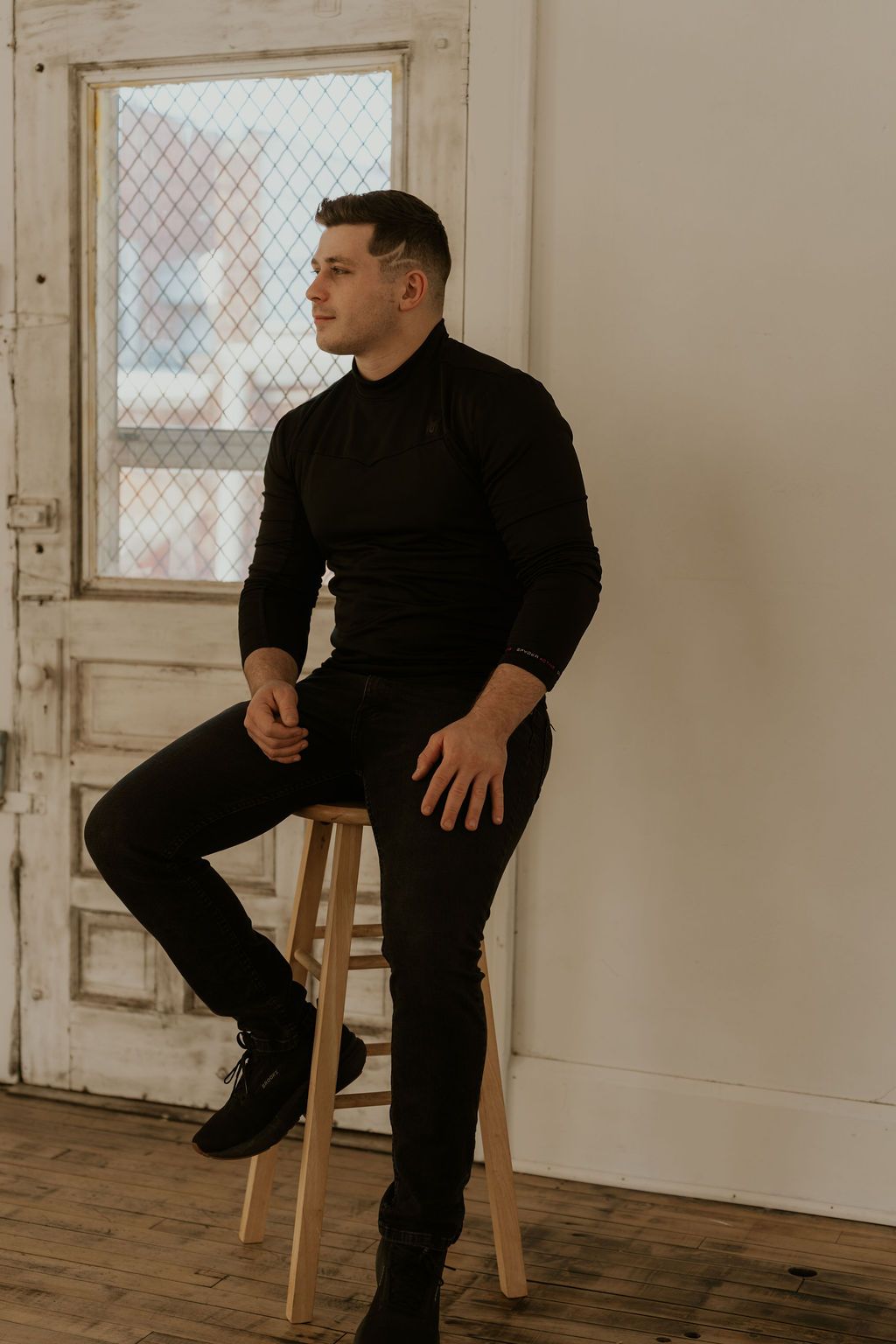 Man in all black outfit sitting on stool in white studio space.