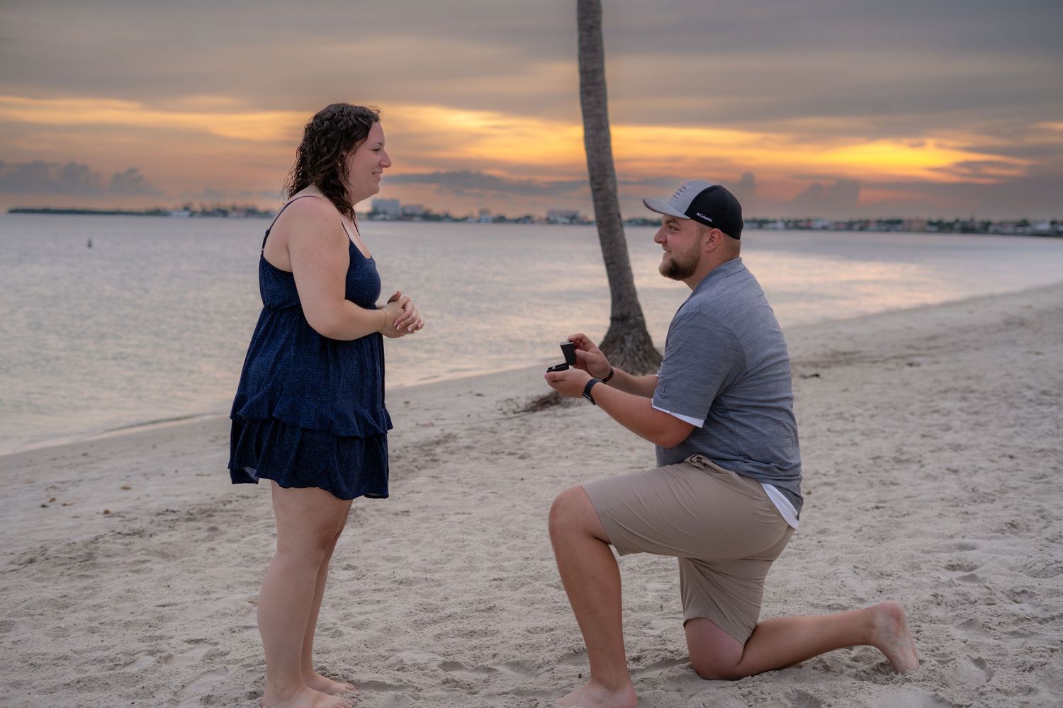 Man kneeling on beach proposing to woman at colorful tropical sunset.