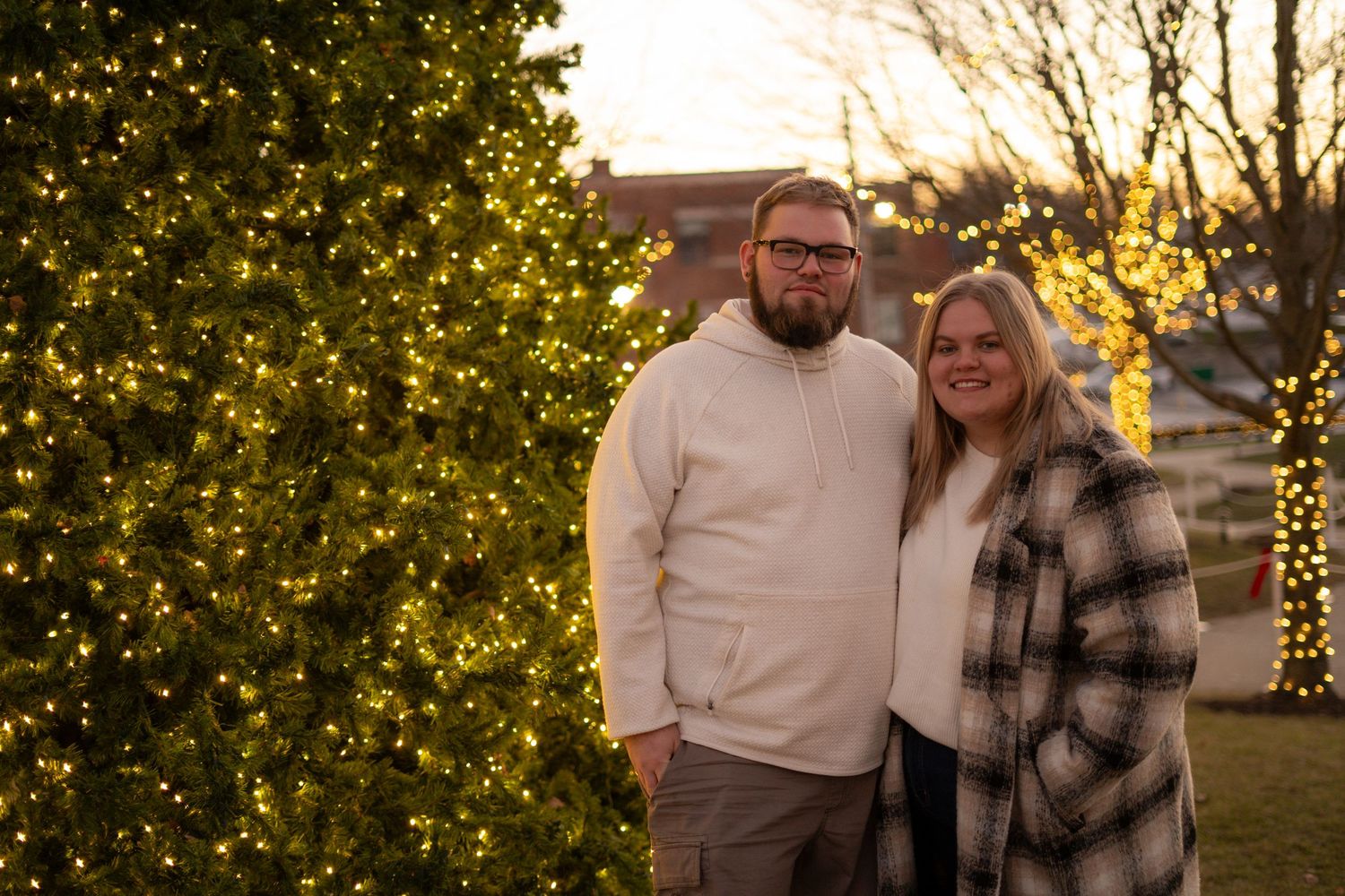 Couple smiling together beside a decorated Christmas tree outdoors at dusk.