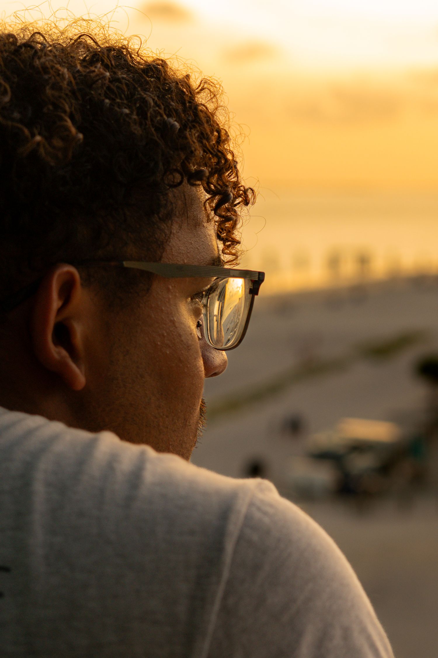 Man with curly hair and glasses gazing at golden beach sunset from above.