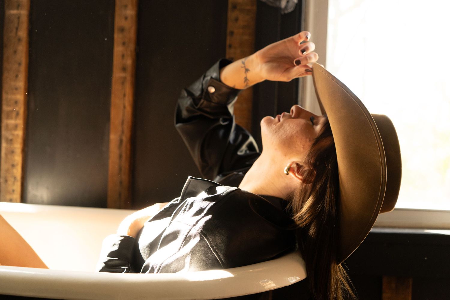 Woman in black outfit and wide-brim hat relaxing in a white bathtub near a sunlit window.