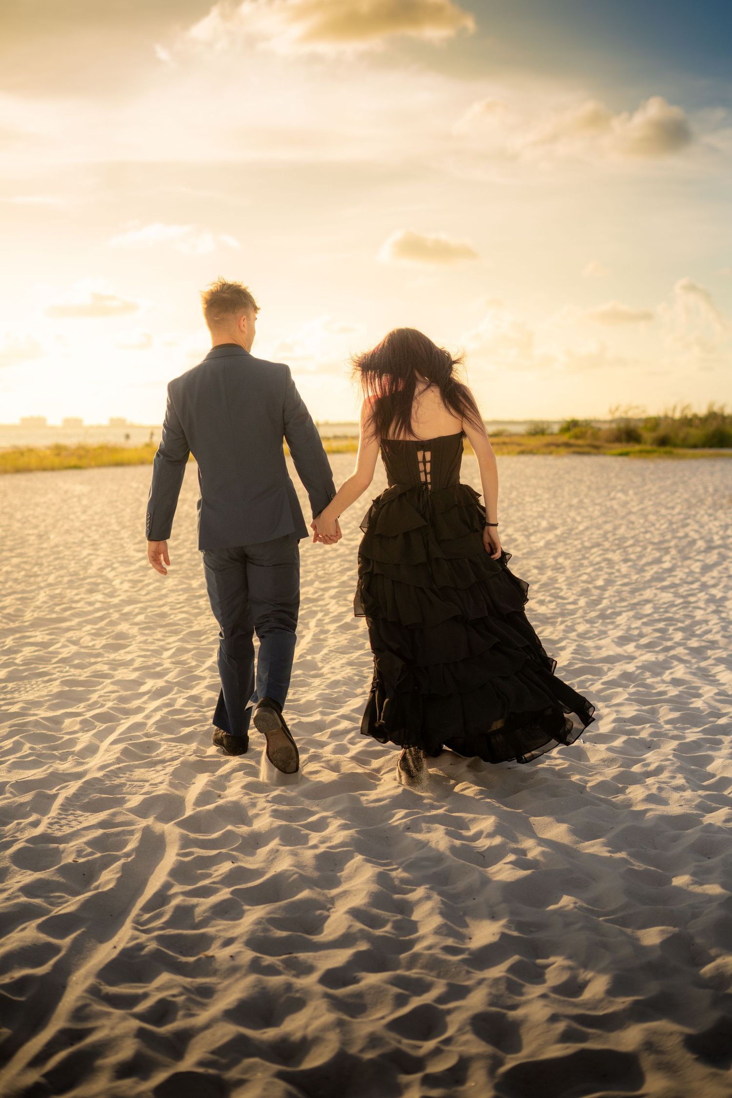Couple in formal wear holding hands walking away on sandy beach at sunset.