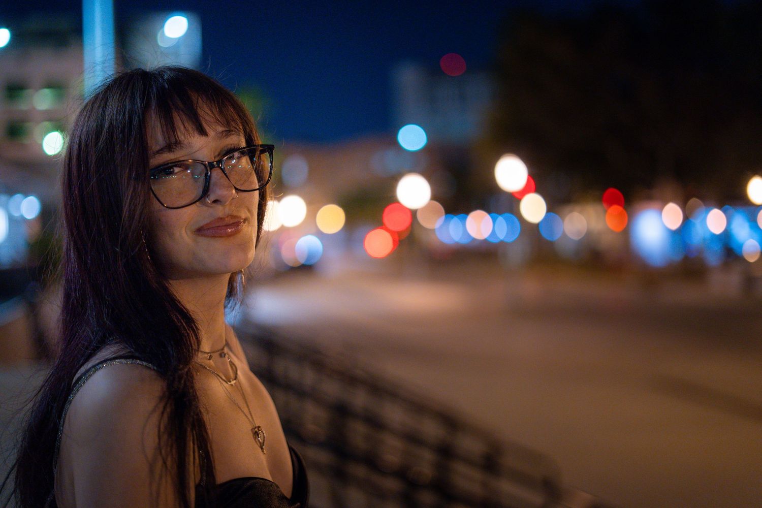 Young woman with glasses posing on city street at night with bokeh lights.