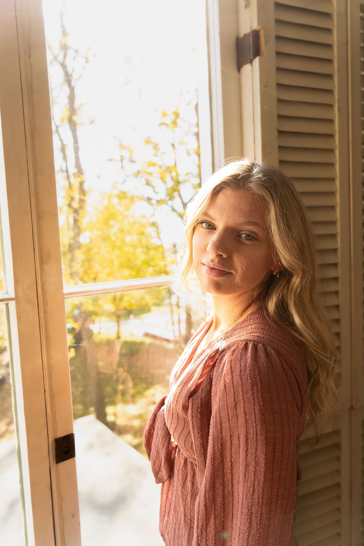Blonde woman in pink sweater standing by sunlit window with autumn view.