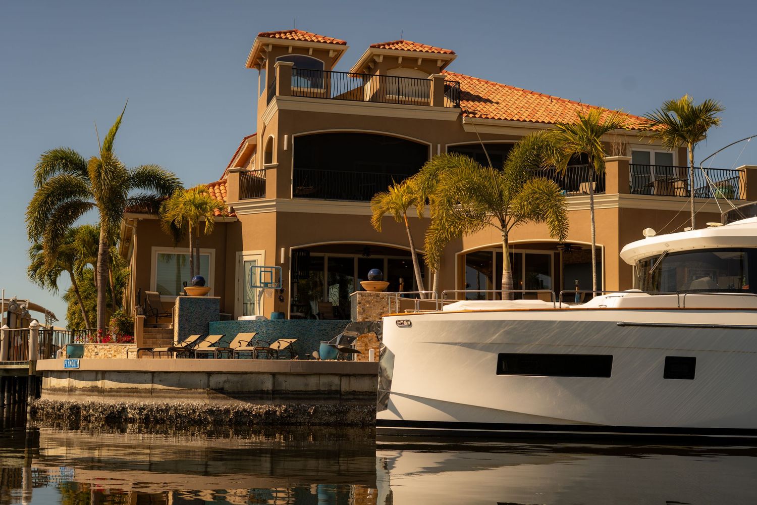 Luxury waterfront mansion with yacht docked at private pier surrounded by palm trees.