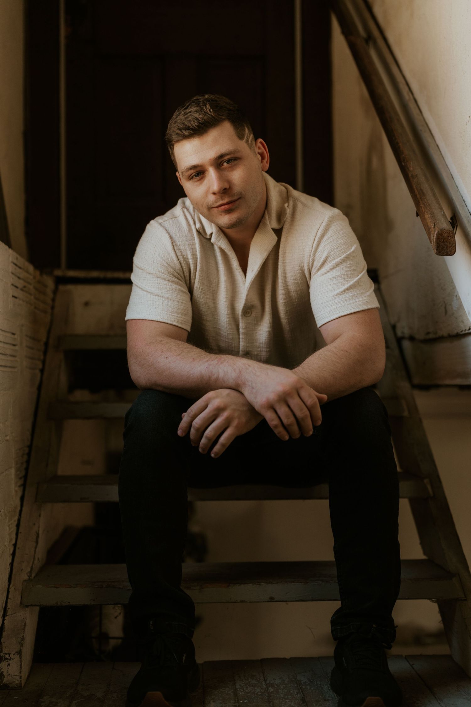 Muscular young man in white polo shirt sitting relaxed on old wooden stairs in dark setting.