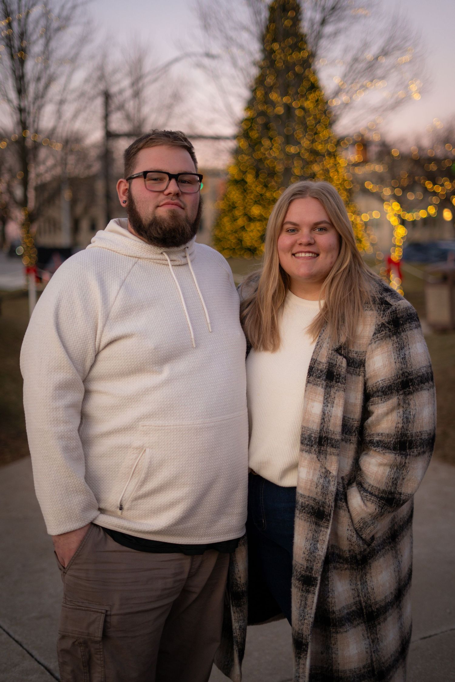 Couple posing together outdoors near Christmas tree lights at dusk.