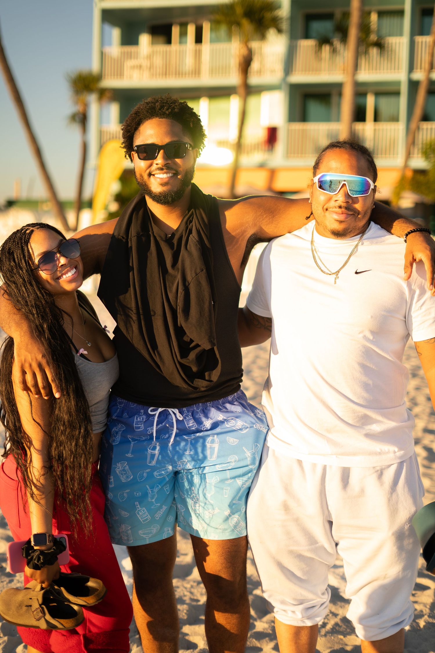 Three friends smiling and posing together on a sunny beach, wearing sunglasses and summer outfits.
