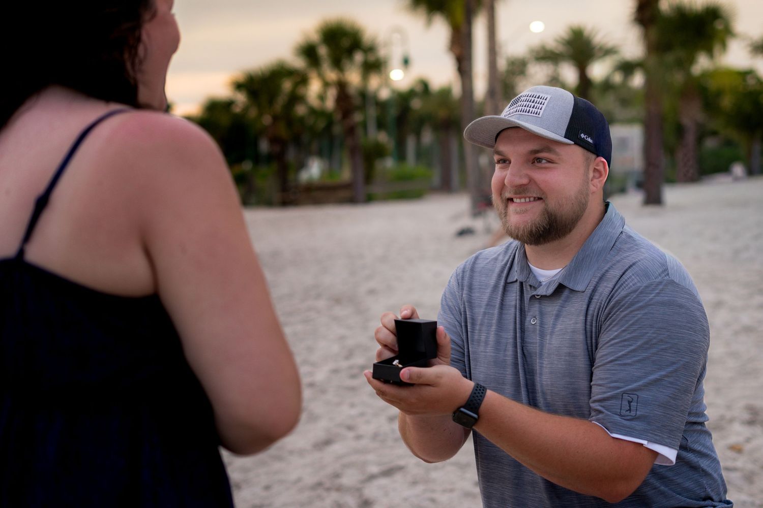 Man proposing on beach at sunset holding open ring box.