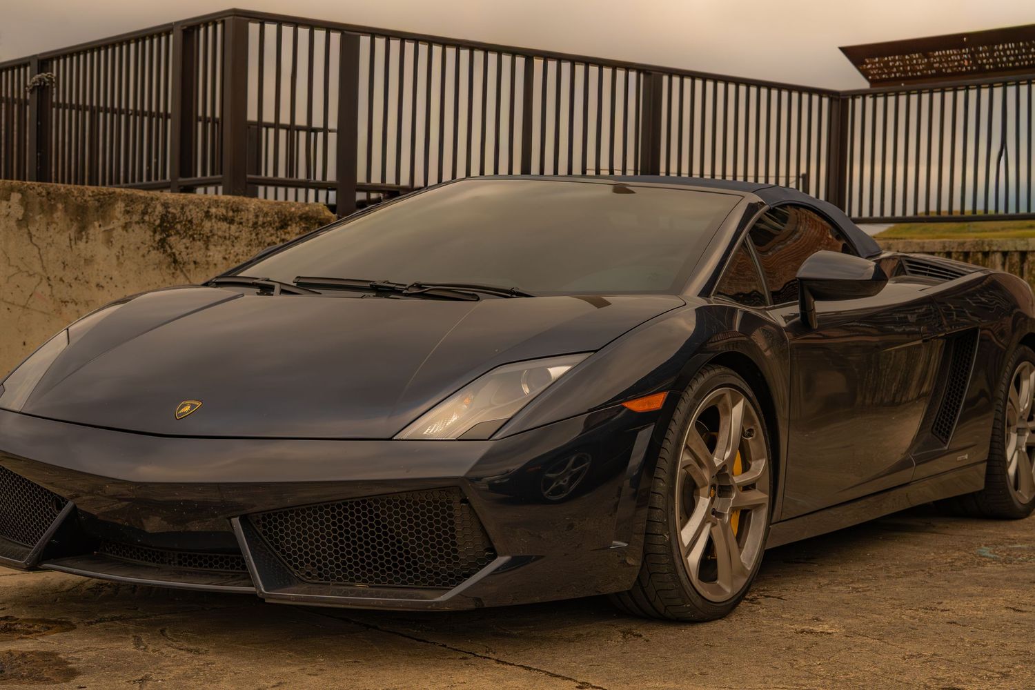 A sleek black Lamborghini Gallardo sports car parked on a sandy surface under an overcast sky.