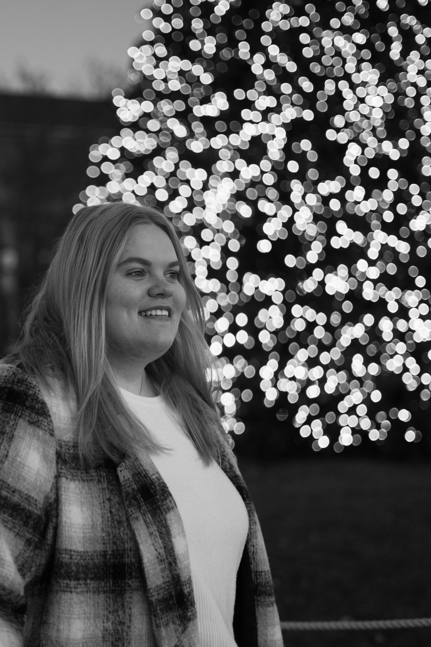 Smiling woman in plaid jacket stands before bokeh Christmas tree lights, black and white photo.