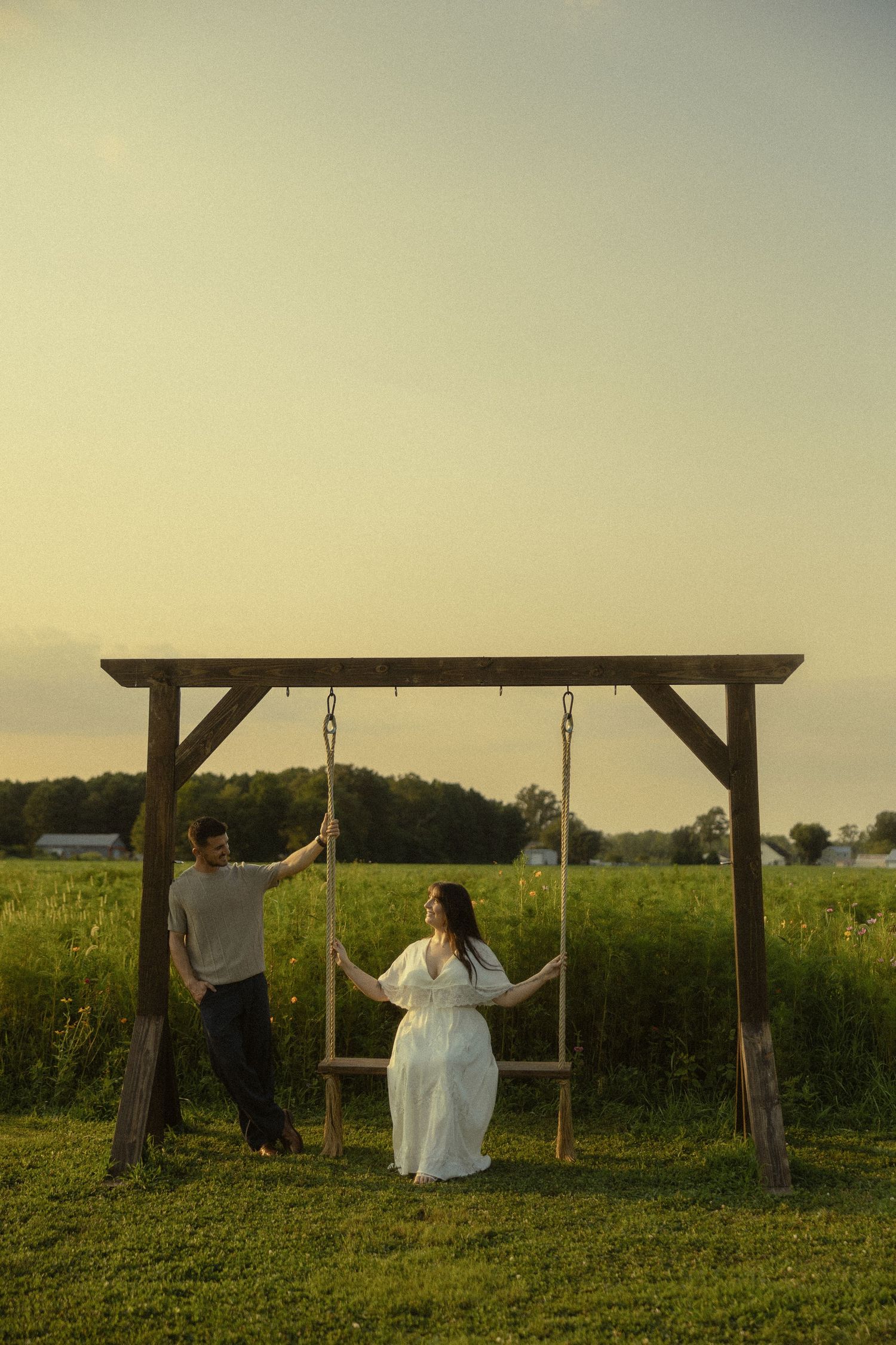 Couple at rustic wooden swing set in green field at golden hour, woman in white dress, romantic outdoor maternity photo.