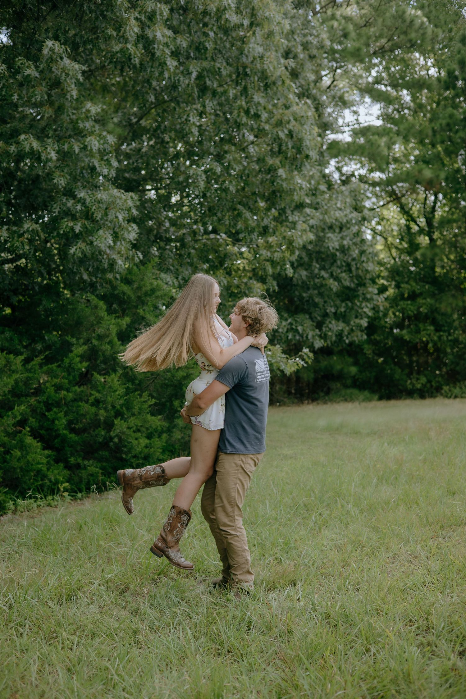 A playful couple spins together in a grassy field surrounded by trees.