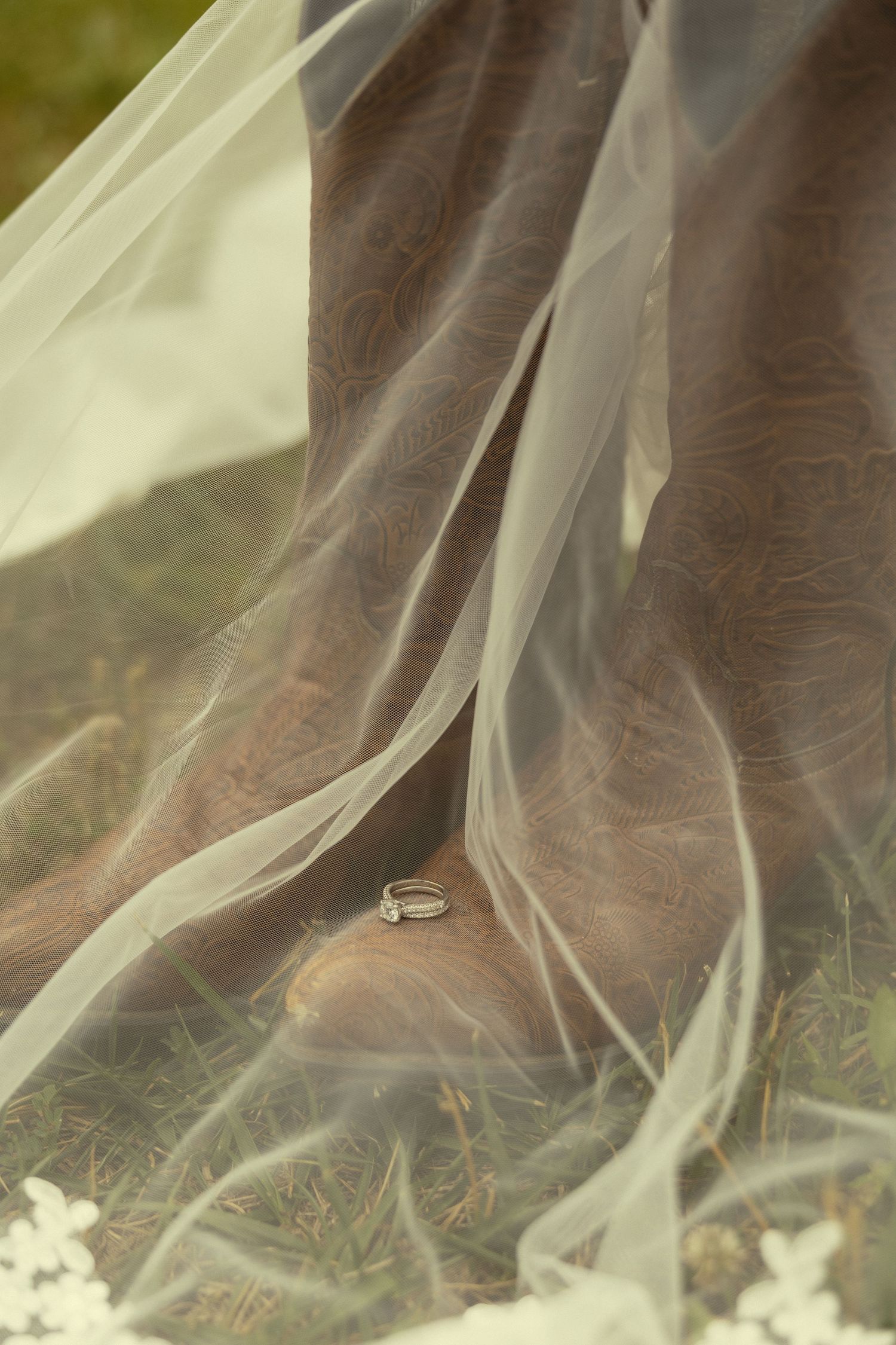 Wedding rings resting on brown cowboy boots draped with a flowing white bridal veil on green grass.