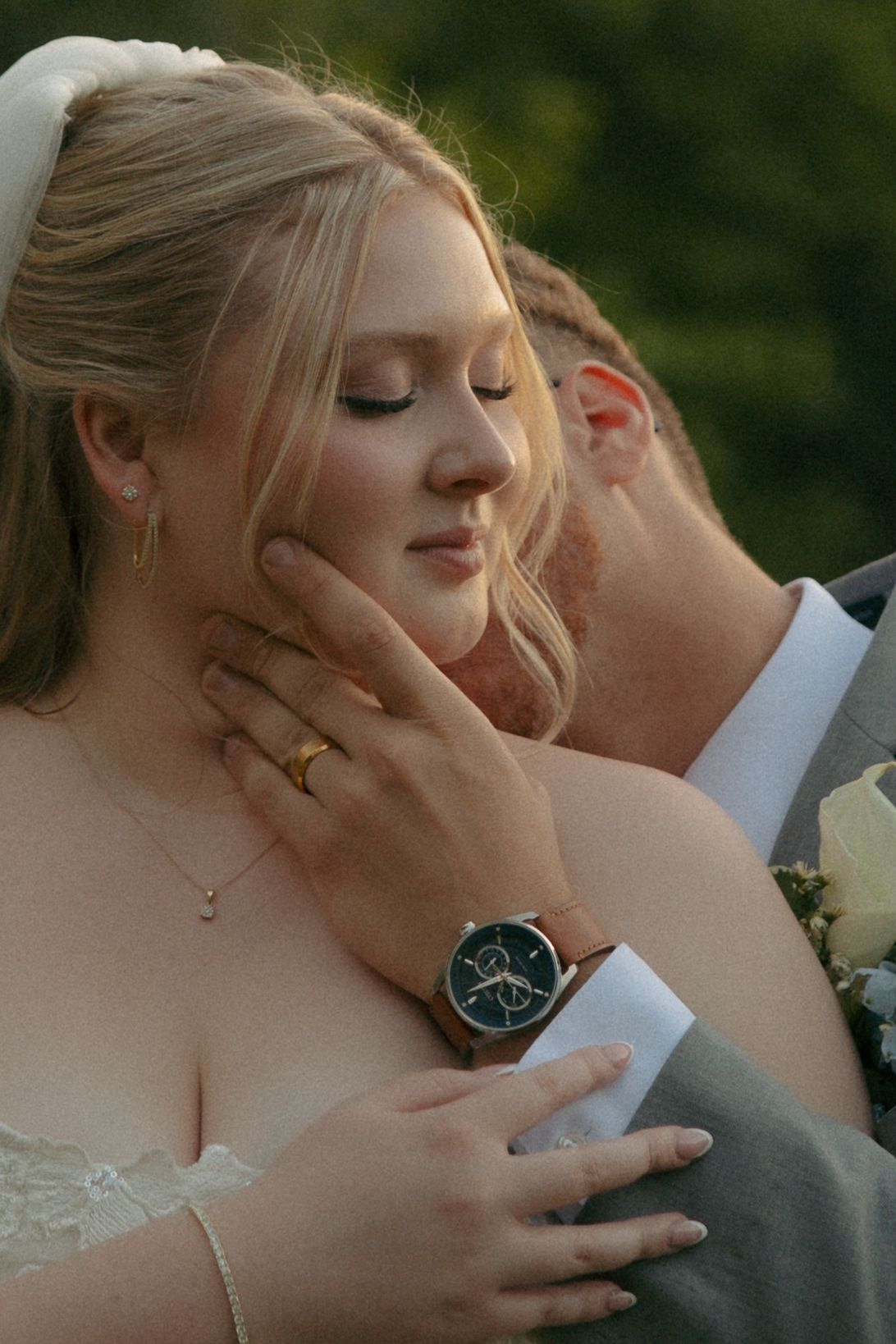A groom tenderly holds his bride's face during their wedding, she wears a gold ring and he sports a brown watch.