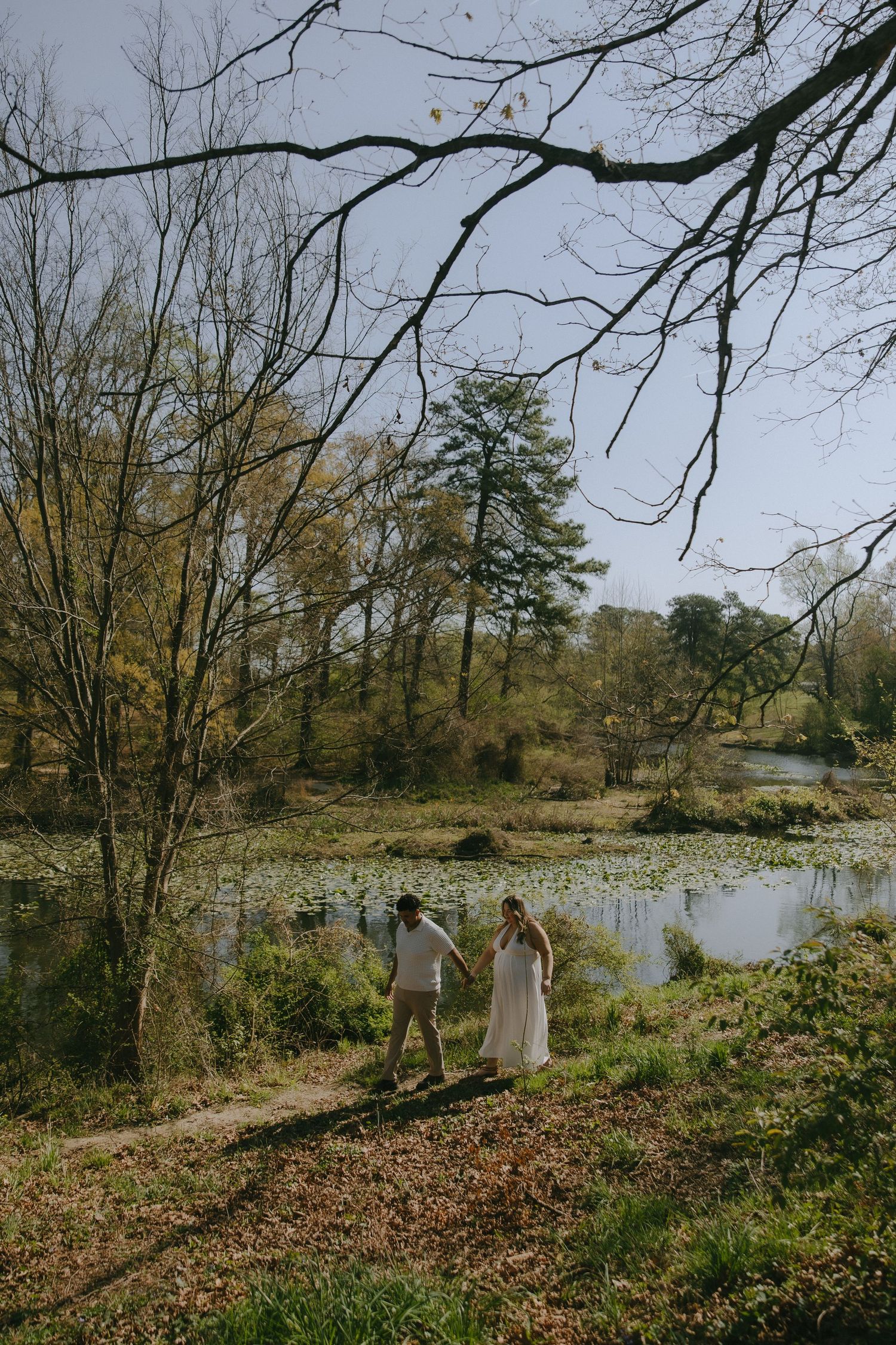 Couple in white wedding attire holding hands while walking along a scenic pond surrounded by autumn trees.