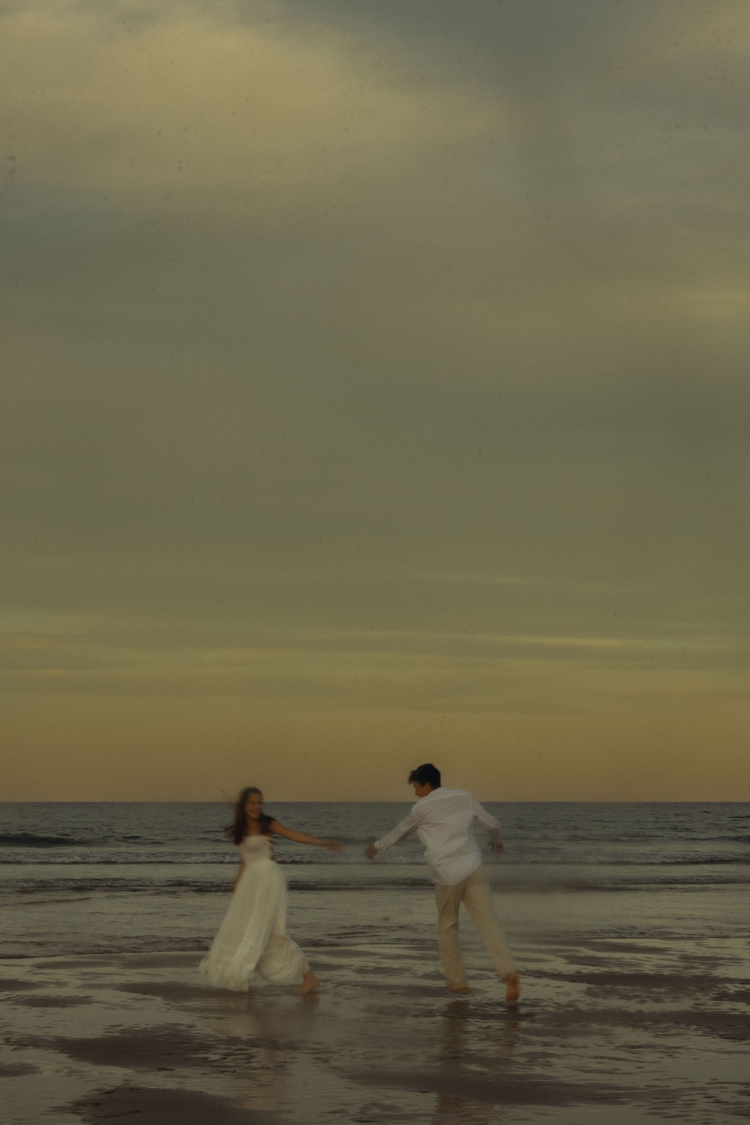 A couple in white attire dancing romantically on a beach at sunset with reflective wet sand.