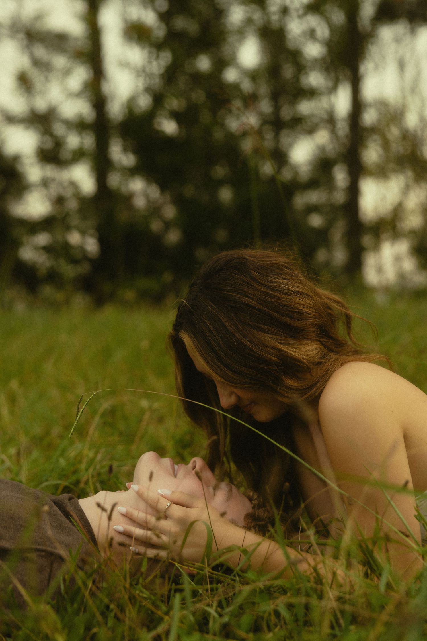 Romantic couple lying in green grass field, woman leaning over man tenderly, trees in soft focus background.