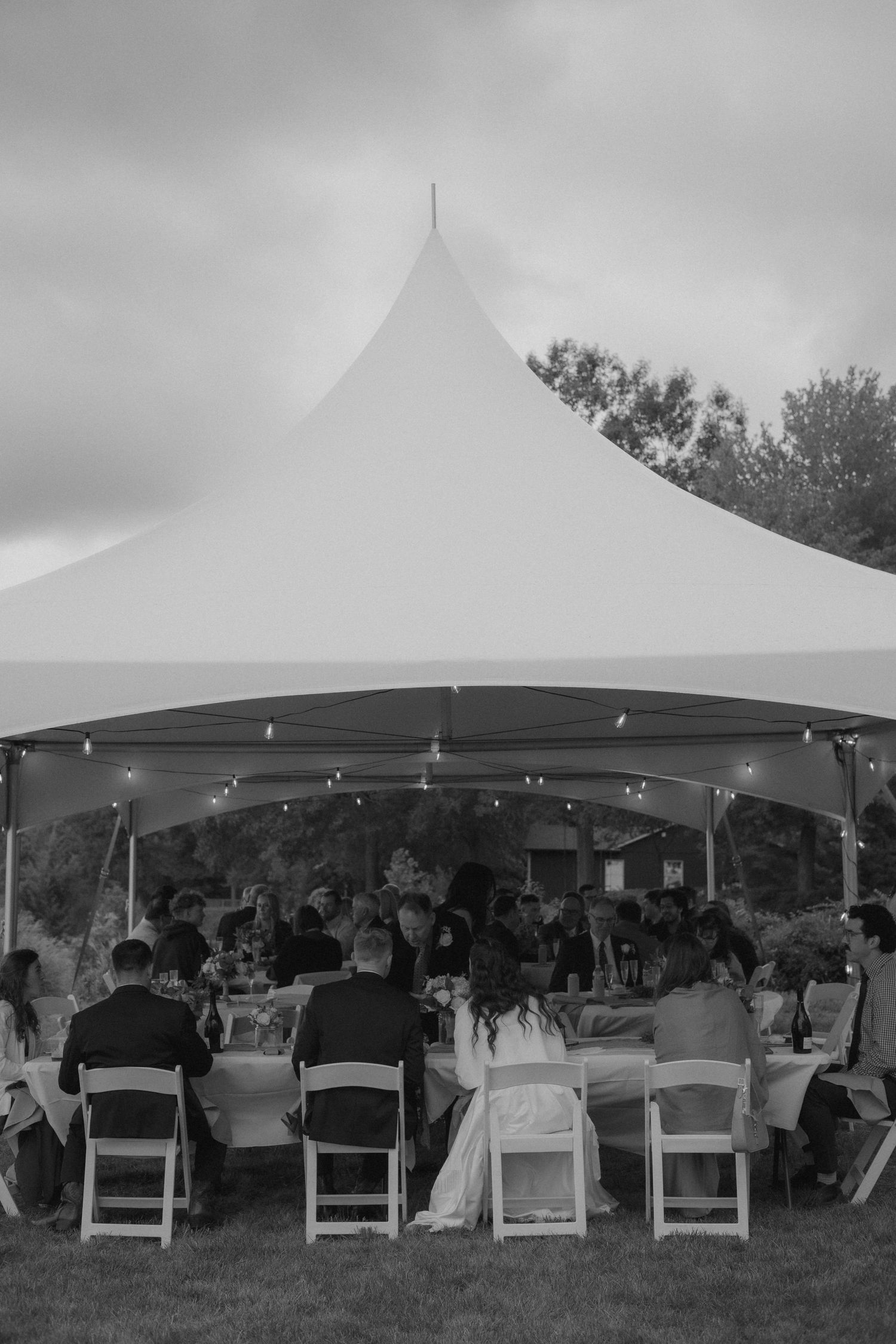 Black and white photo of guests dining under an elegant white tent with string lights at an outdoor wedding reception.