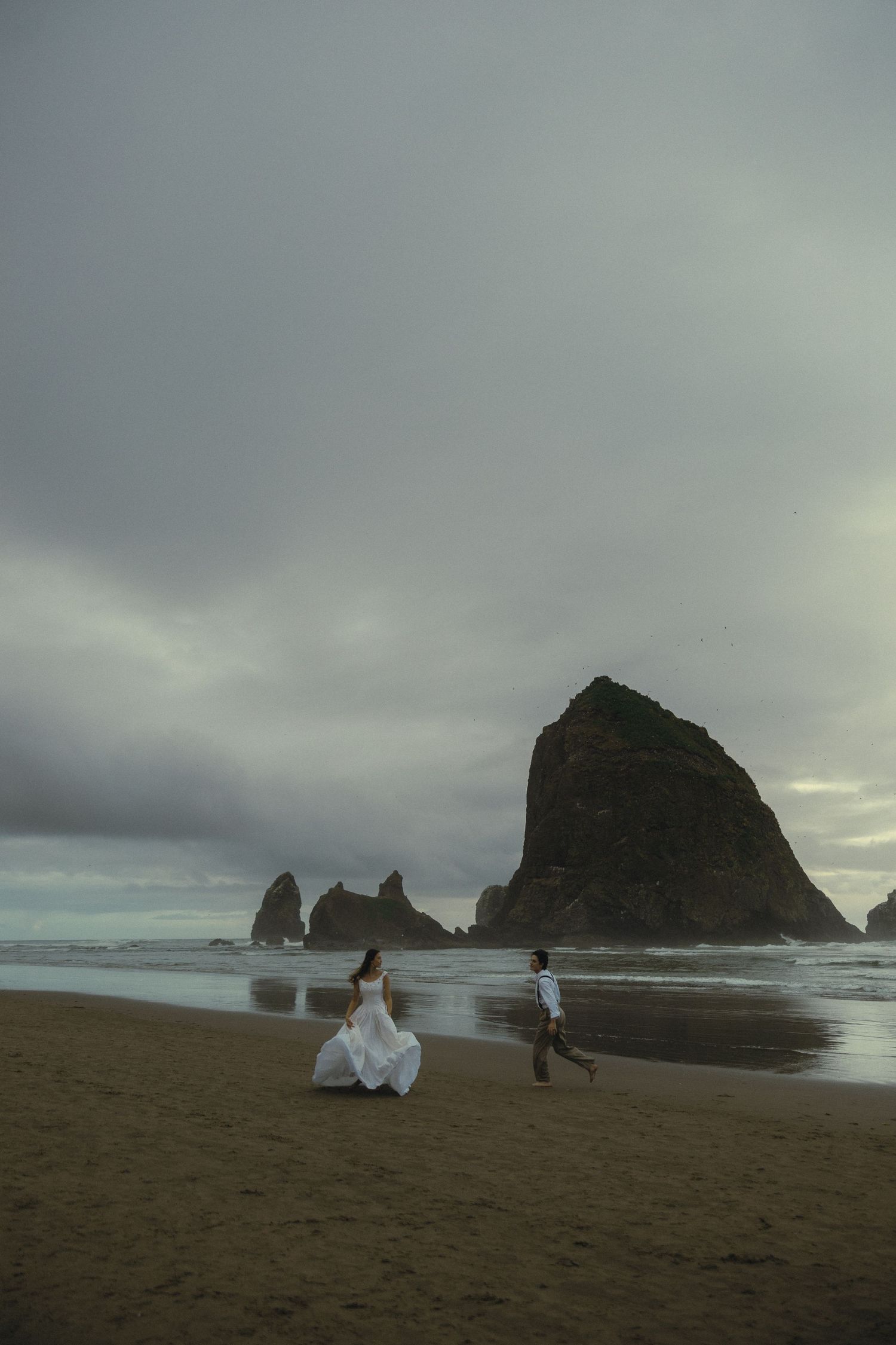 Bride and groom walking on a dramatic beach with iconic sea stack rock formation under cloudy skies.