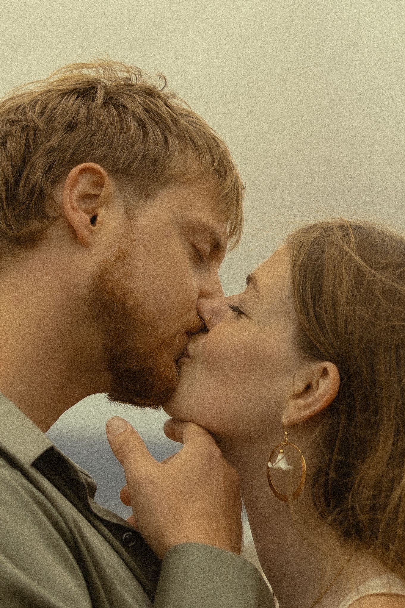 A couple shares a tender kiss, the man with a red beard gently holding the woman's hands near their lips.