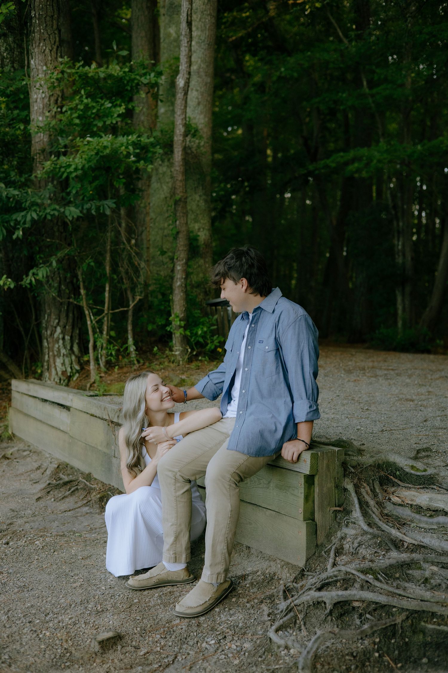 A couple sits on a wooden bench in a lush forest, sharing a tender moment in casual attire.
