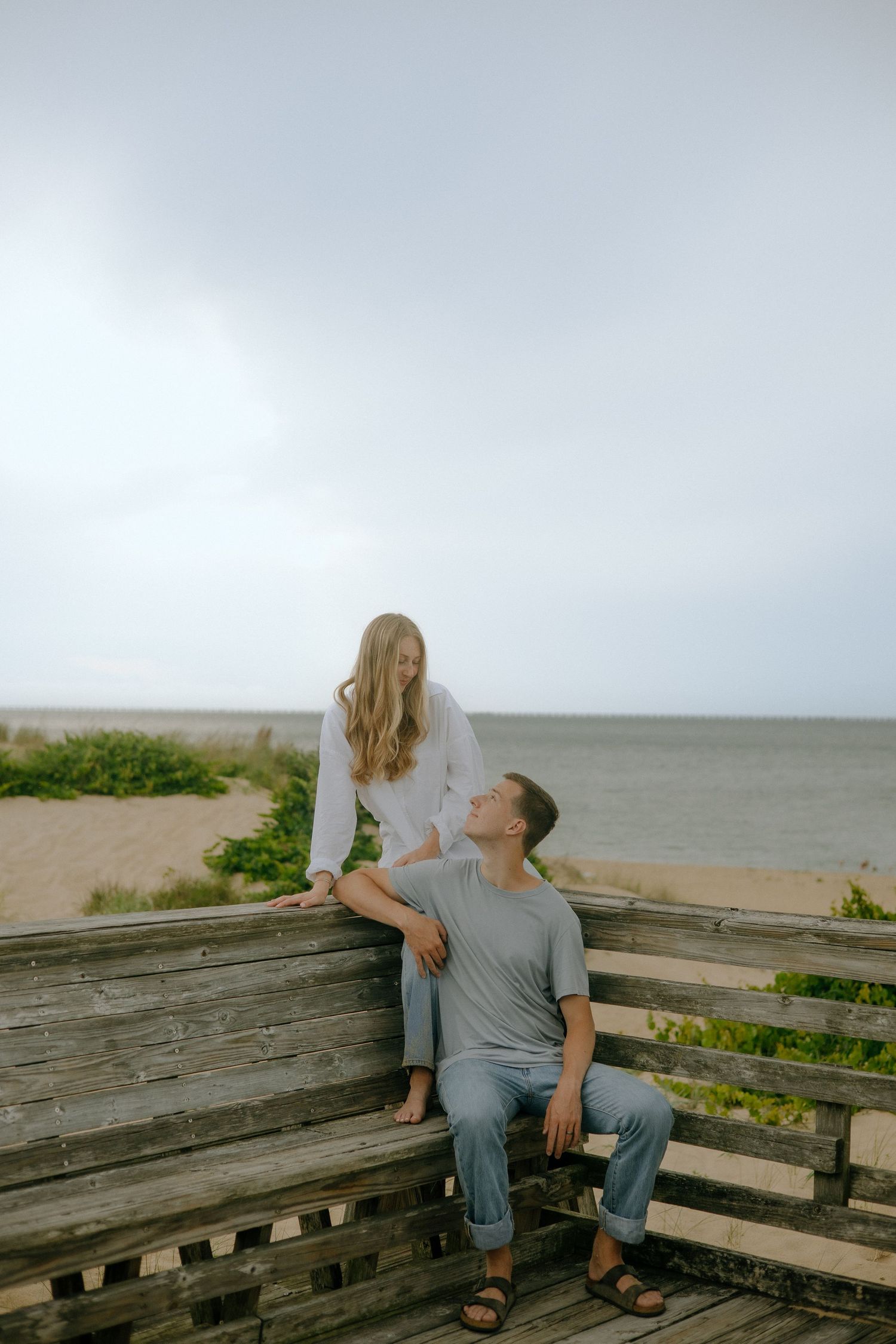 A couple shares a tender moment on a weathered wooden boardwalk by the beach, woman in white standing above seated man.