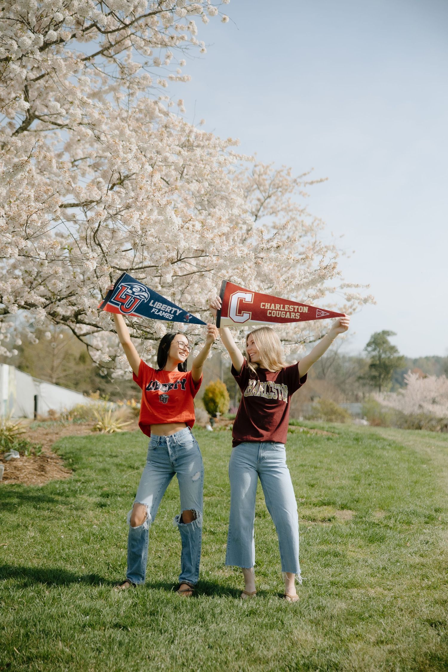 Two girls holding Liberty Flames and Charleston Cougars pennants outdoors near a blooming white cherry blossom tree.
