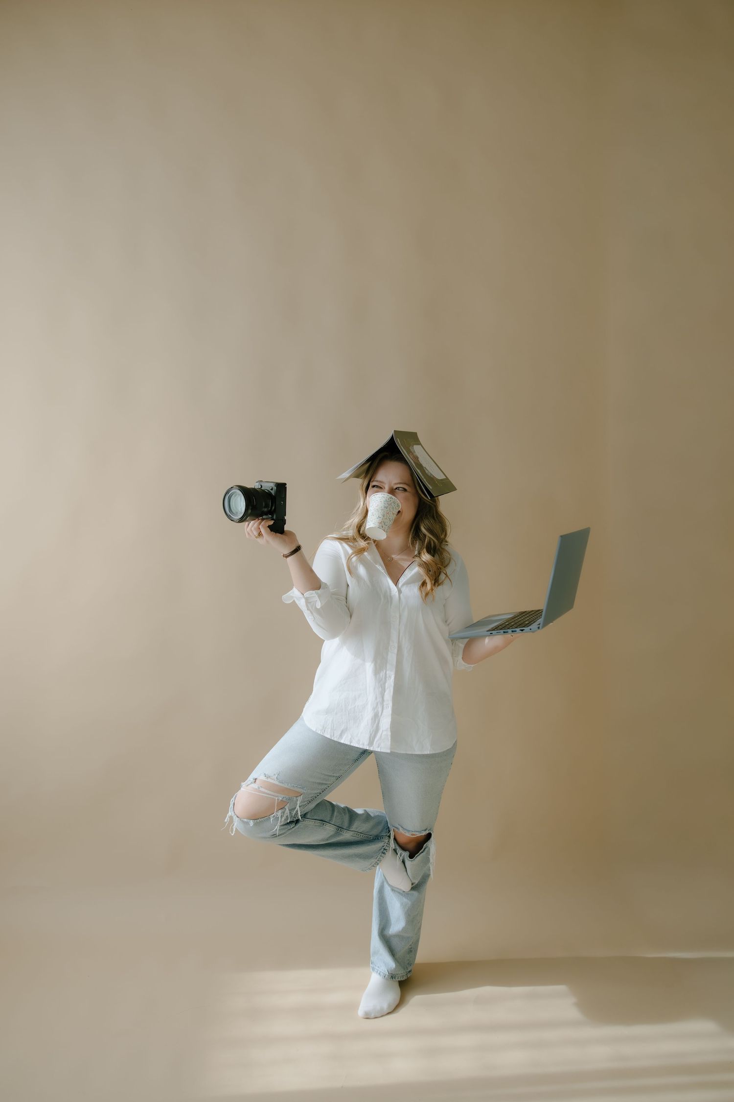 Woman in white shirt and ripped jeans balancing on one leg, holding a camera and laptop while wearing a face mask.