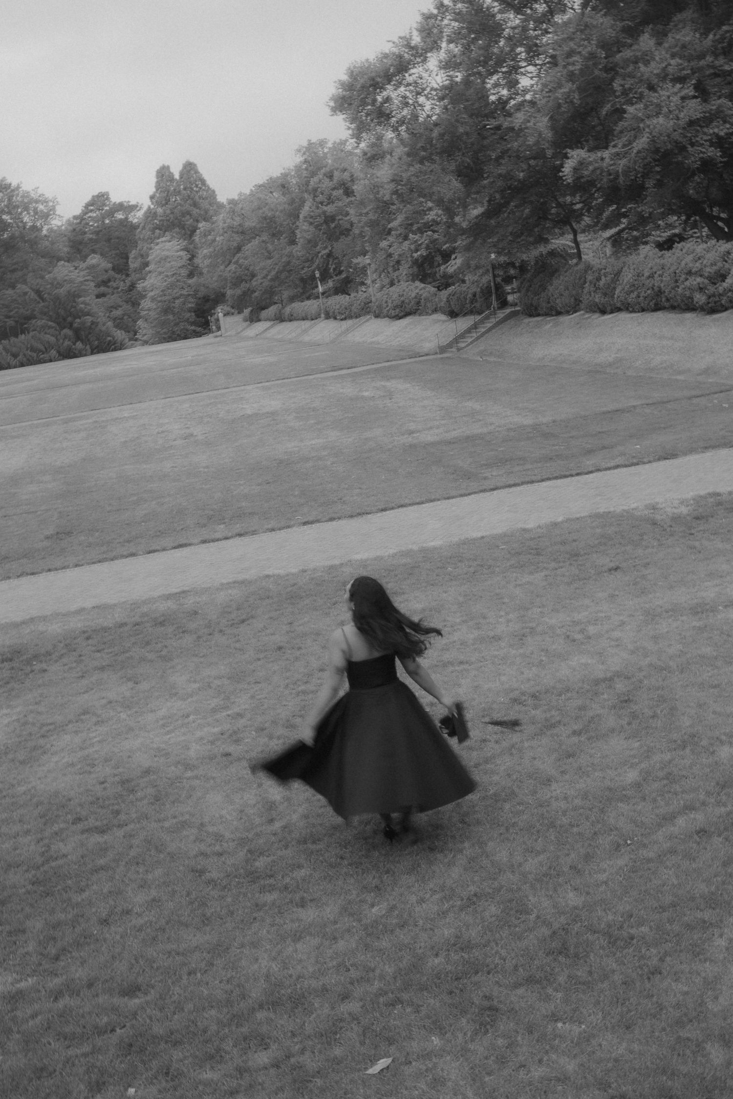 Black and white photo of a woman in a flowing dark dress spinning in a lush green park surrounded by trees.