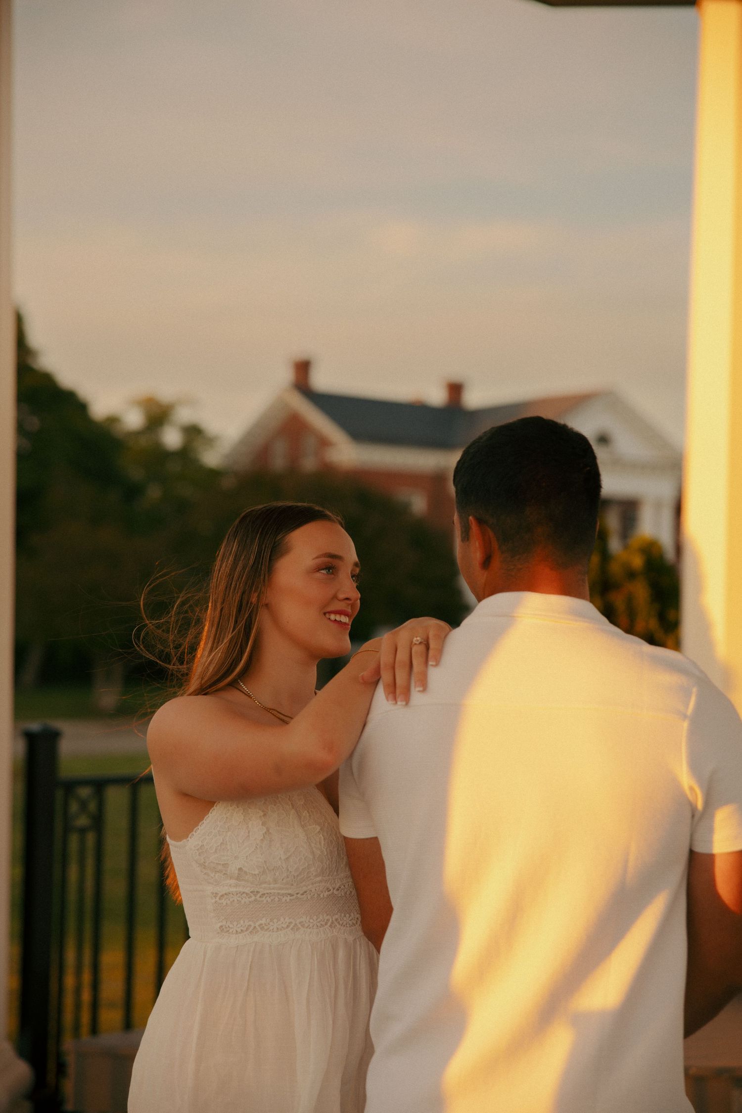 Couple dancing at golden hour sunset, woman in white dress smiling at man in white shirt on outdoor porch.
