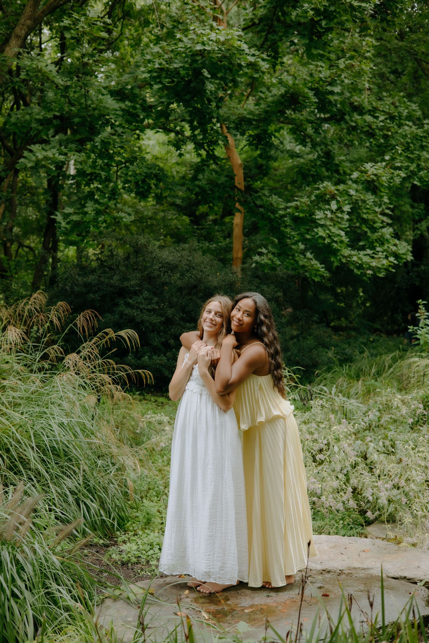 Friends embrace while wearing flowing white and cream dresses in a garden setting.