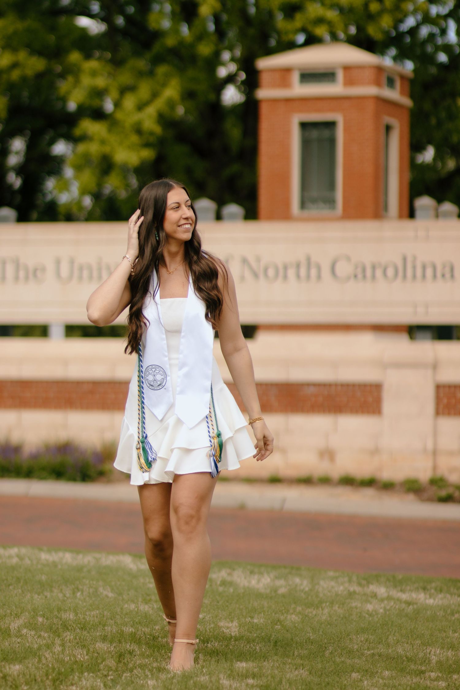 A person in a white outfit poses on a grassy lawn in front of a university building.