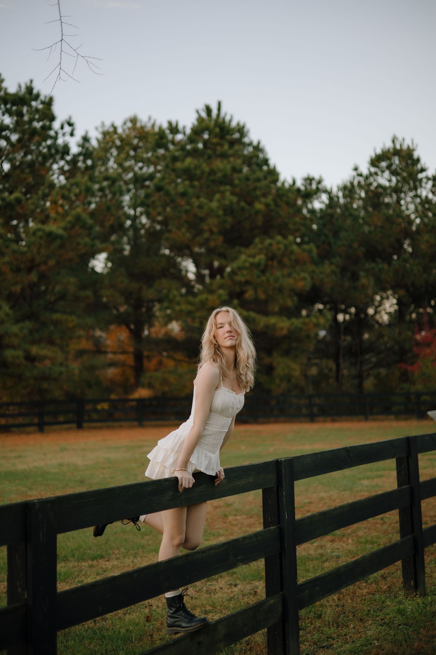 A person in a white dress stands balanced on a black wooden fence with autumn trees in the background.
