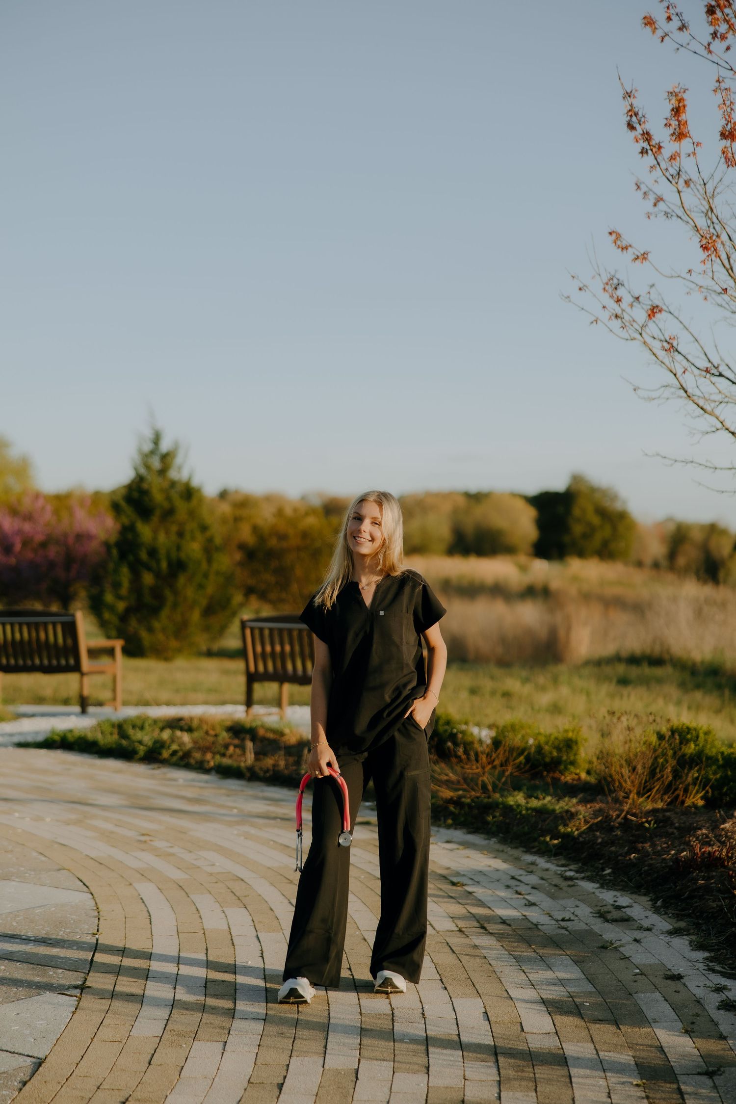 Young woman in black scrubs walking on a brick path in a sunny park, carrying a bag, with trees in the background.
