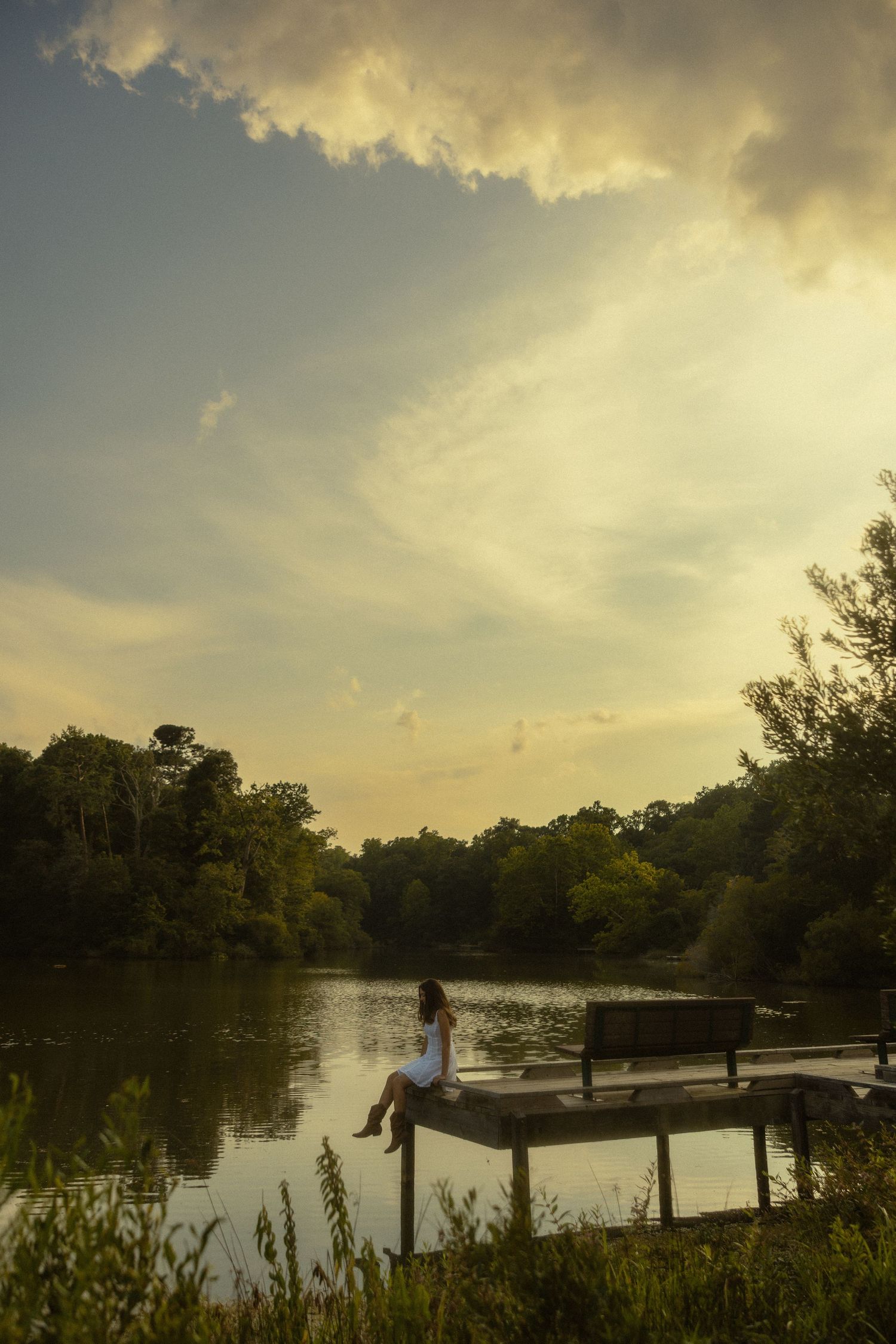 Woman in white dress sits on a dock by a serene lake surrounded by lush trees under a dramatic golden sunset sky.