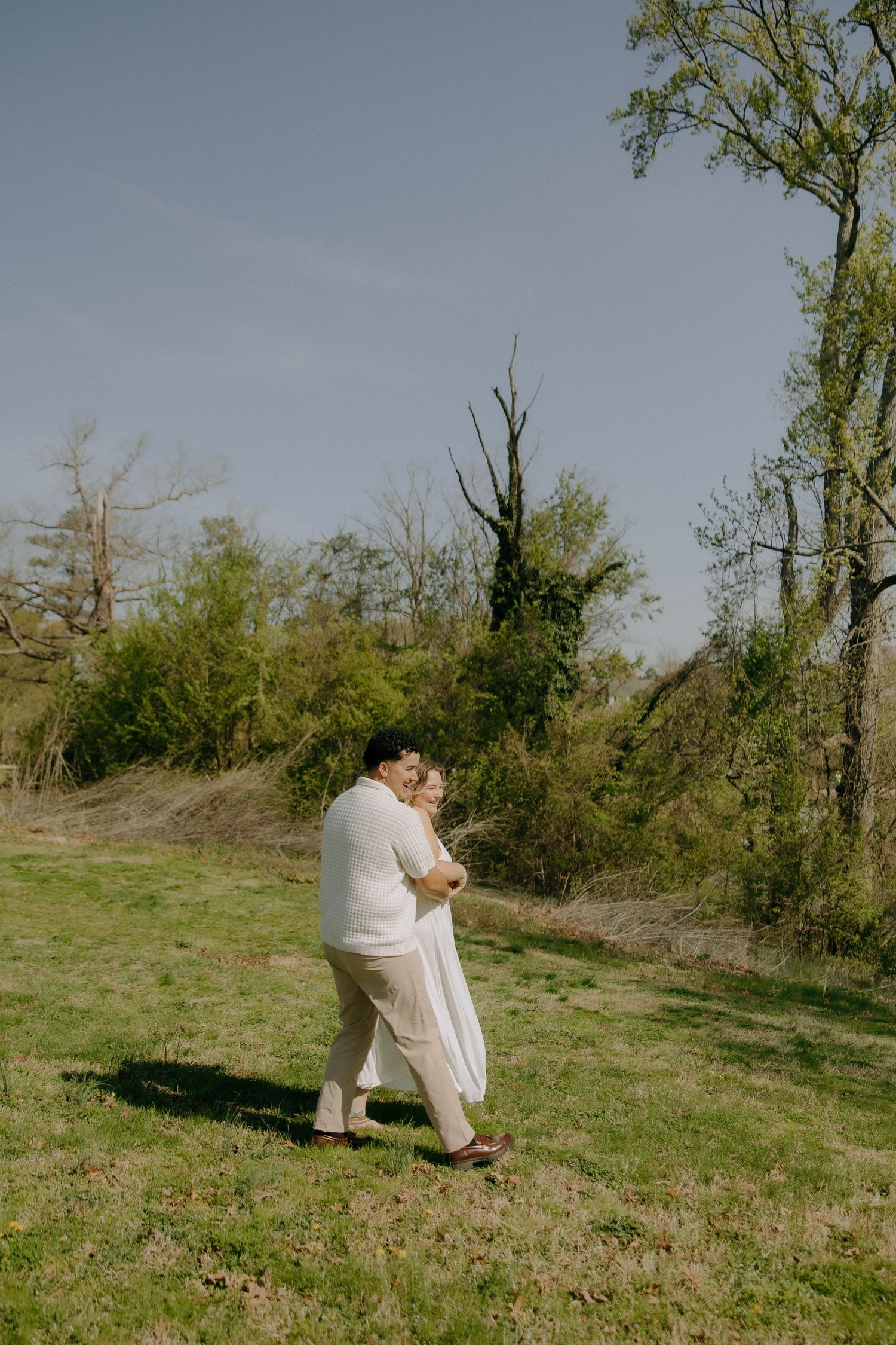 Couple in matching white outfits embracing and dancing on a green meadow with trees in the background.
