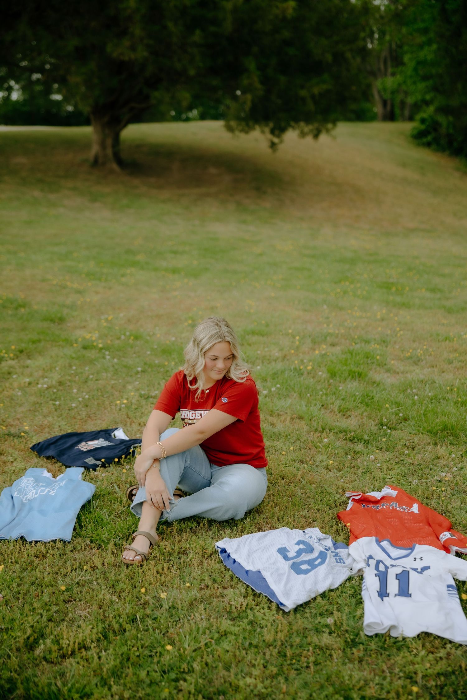 Woman in red shirt sitting on grass surrounded by sports jerseys in a park setting.
