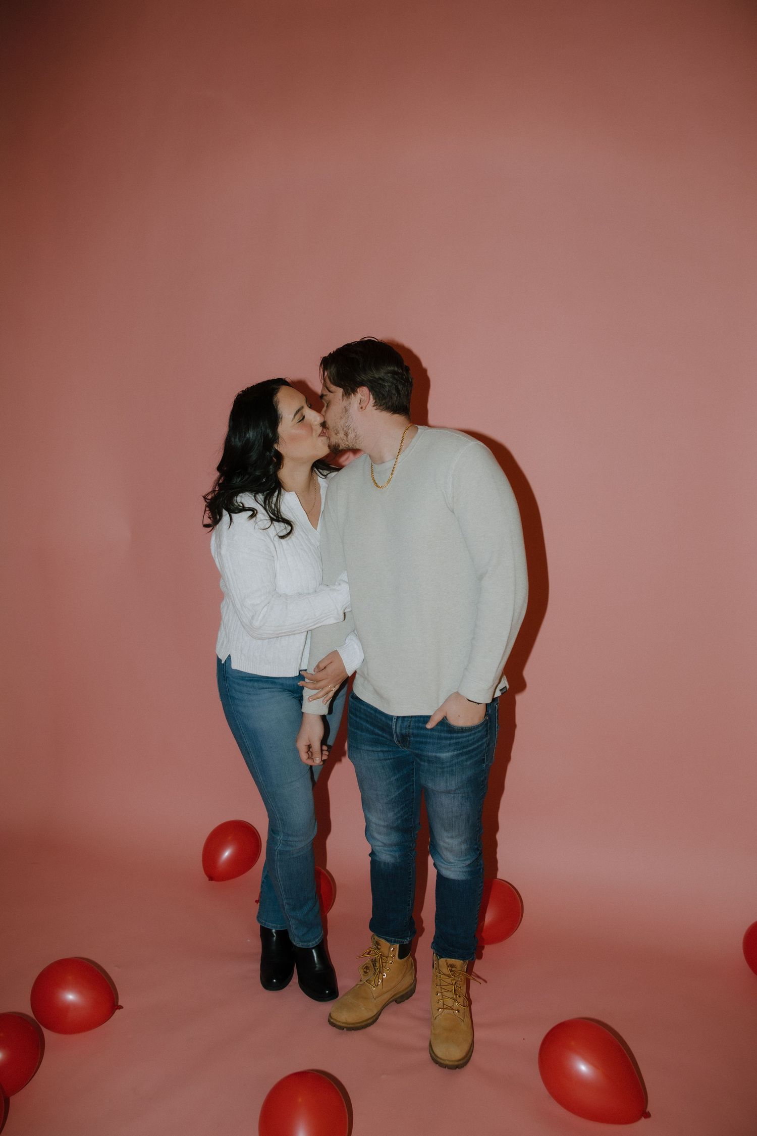 A couple shares a kiss surrounded by red balloons against a pink backdrop, dressed in casual jeans and sweaters.
